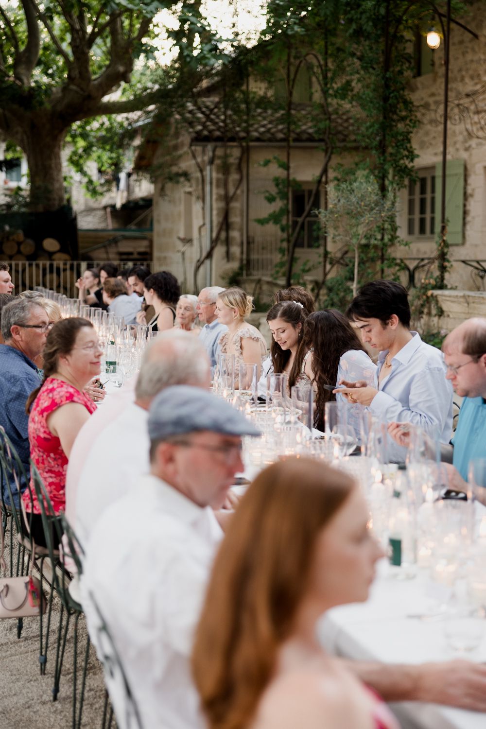 Outdoor wedding reception guests seated at long dining tables under trees with glowing candles at dusk.
