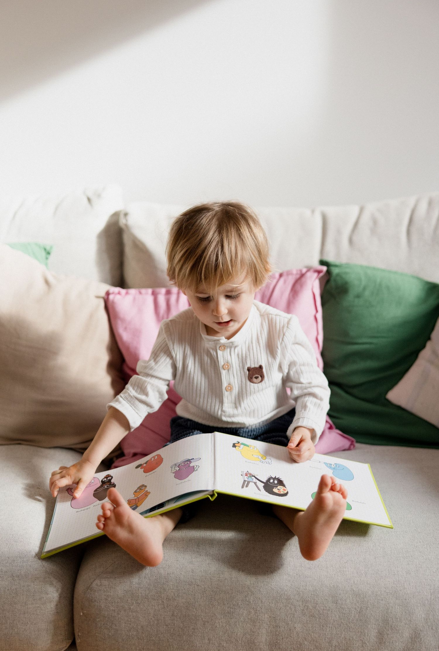 Young toddler sitting barefoot on a couch, reading a colorful picture book with pillows in the background.