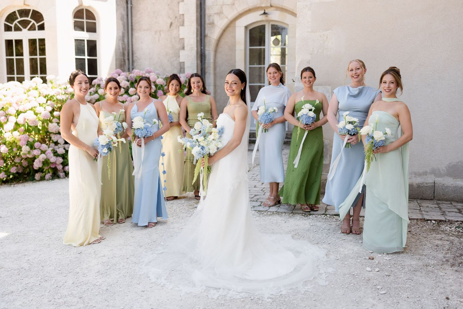 Bride in white gown poses with bridesmaids in mismatched sage green, blue, and yellow dresses holding bouquets outdoors.