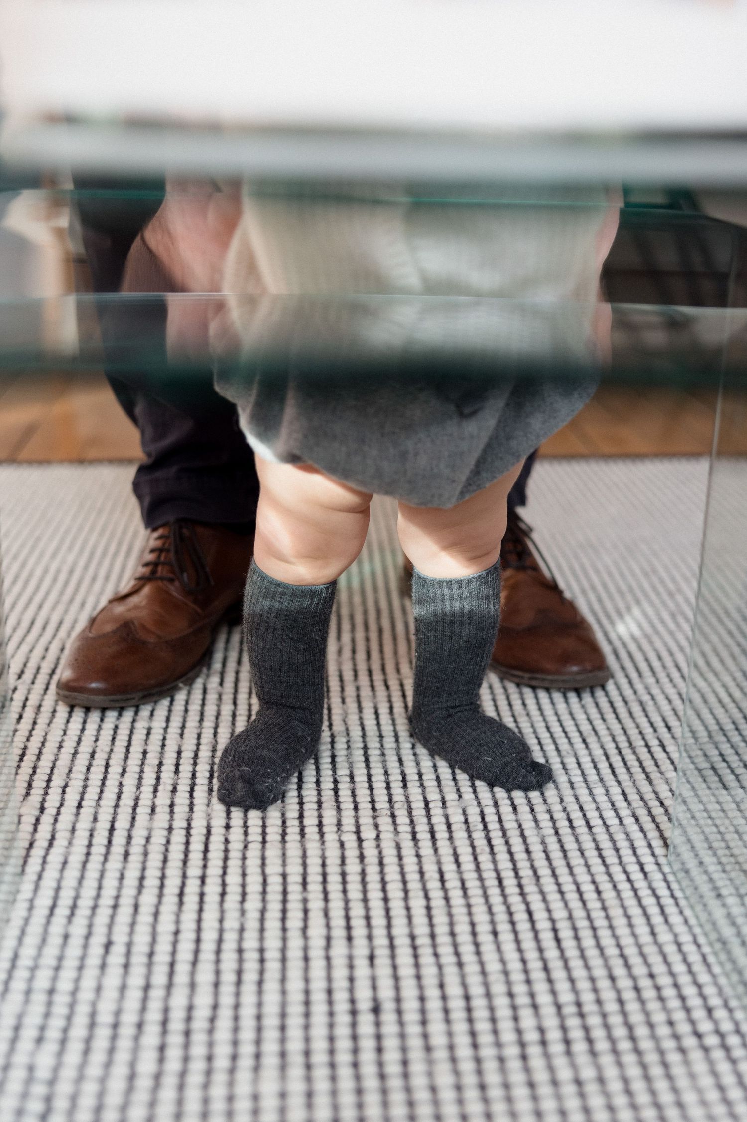 Child in gray knee socks standing between adult legs in brown dress shoes on striped carpet under glass table.