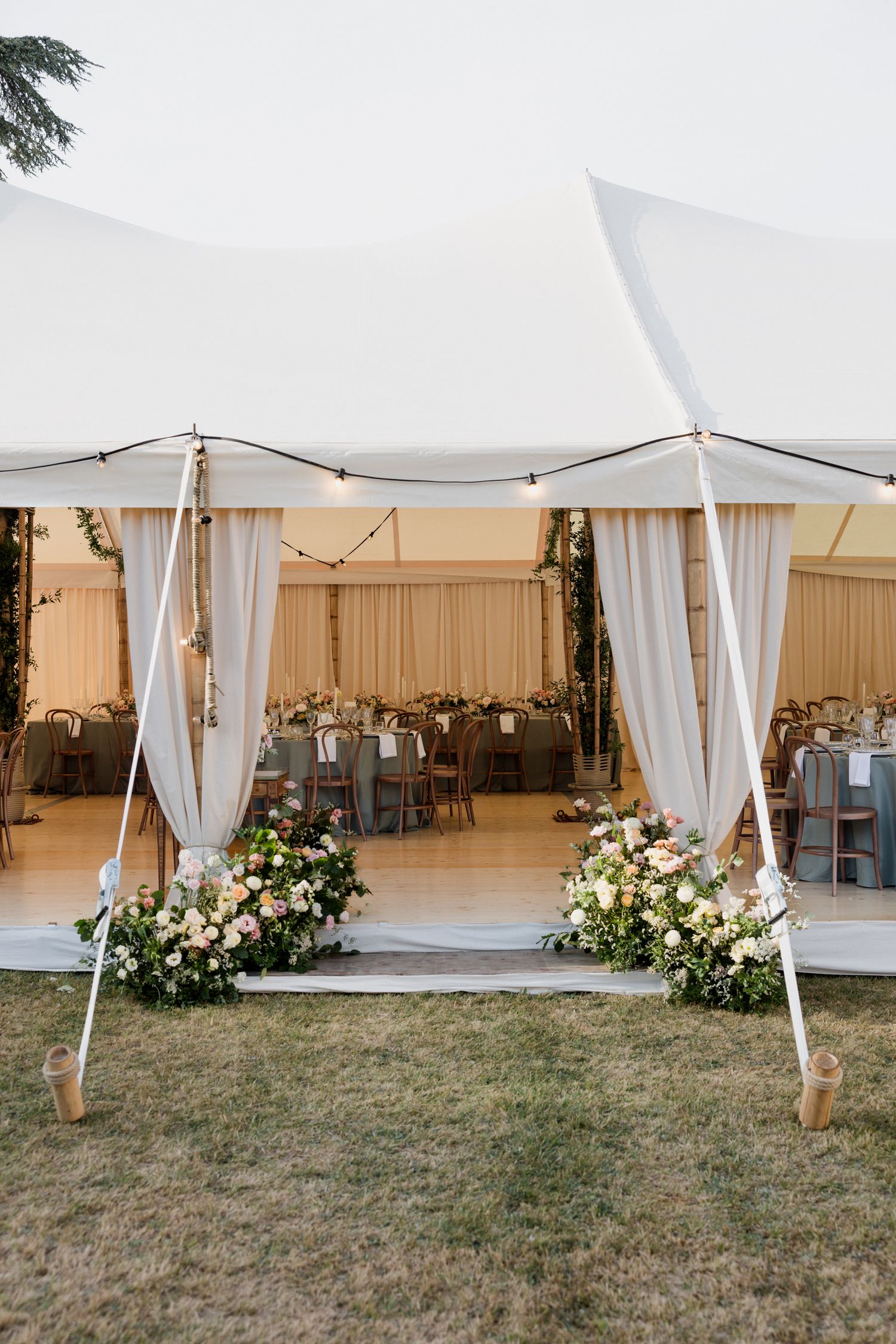 White draped tent canopies with floral arrangements create an elegant outdoor wedding venue setup on a grassy lawn.