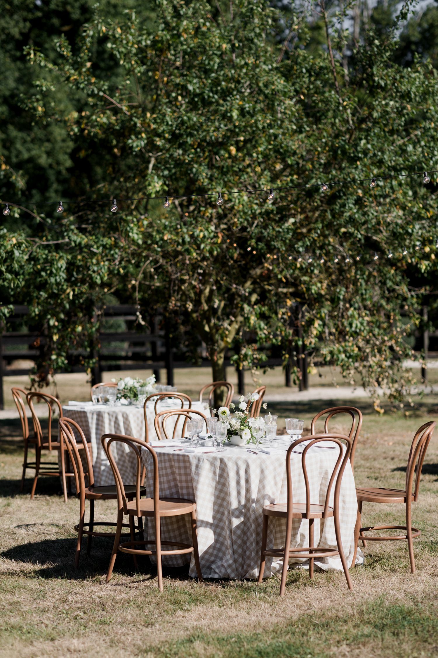 Outdoor wedding reception setup with round tables, white linens and wooden chairs arranged under trees in a garden setting.