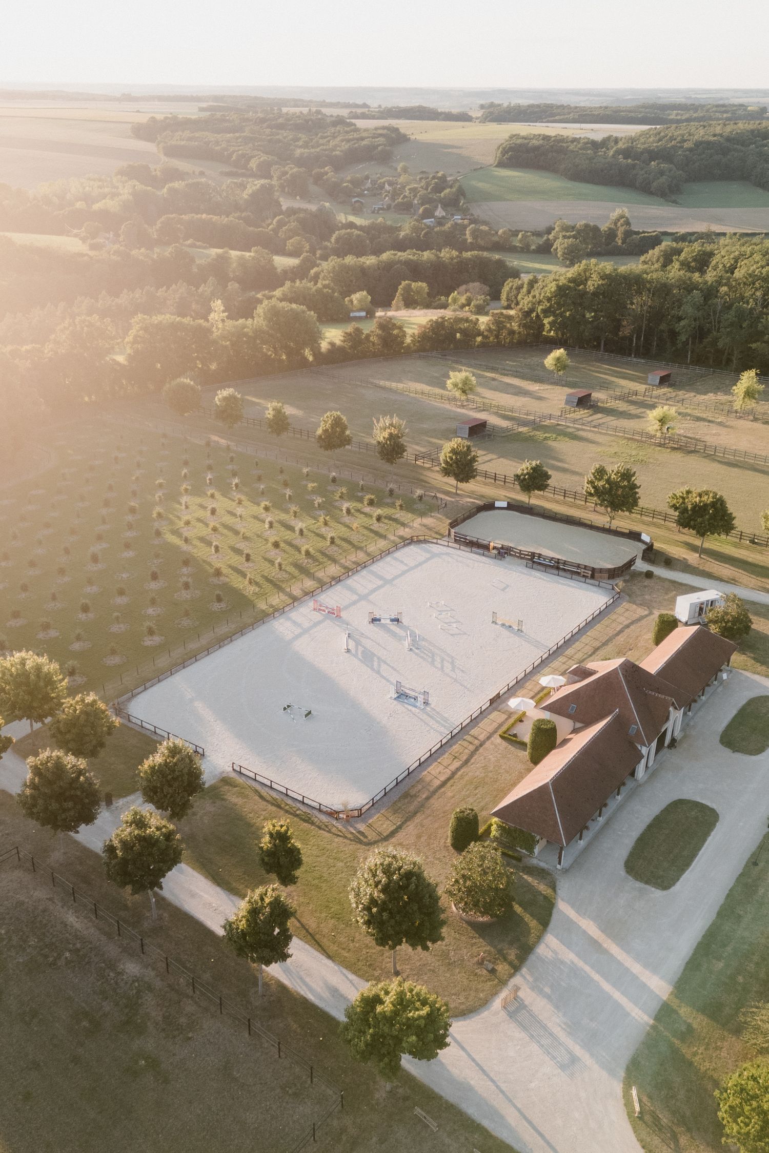 Aerial drone view of a large white equestrian arena with riding facilities surrounded by green countryside at sunset. Photographe mariage To