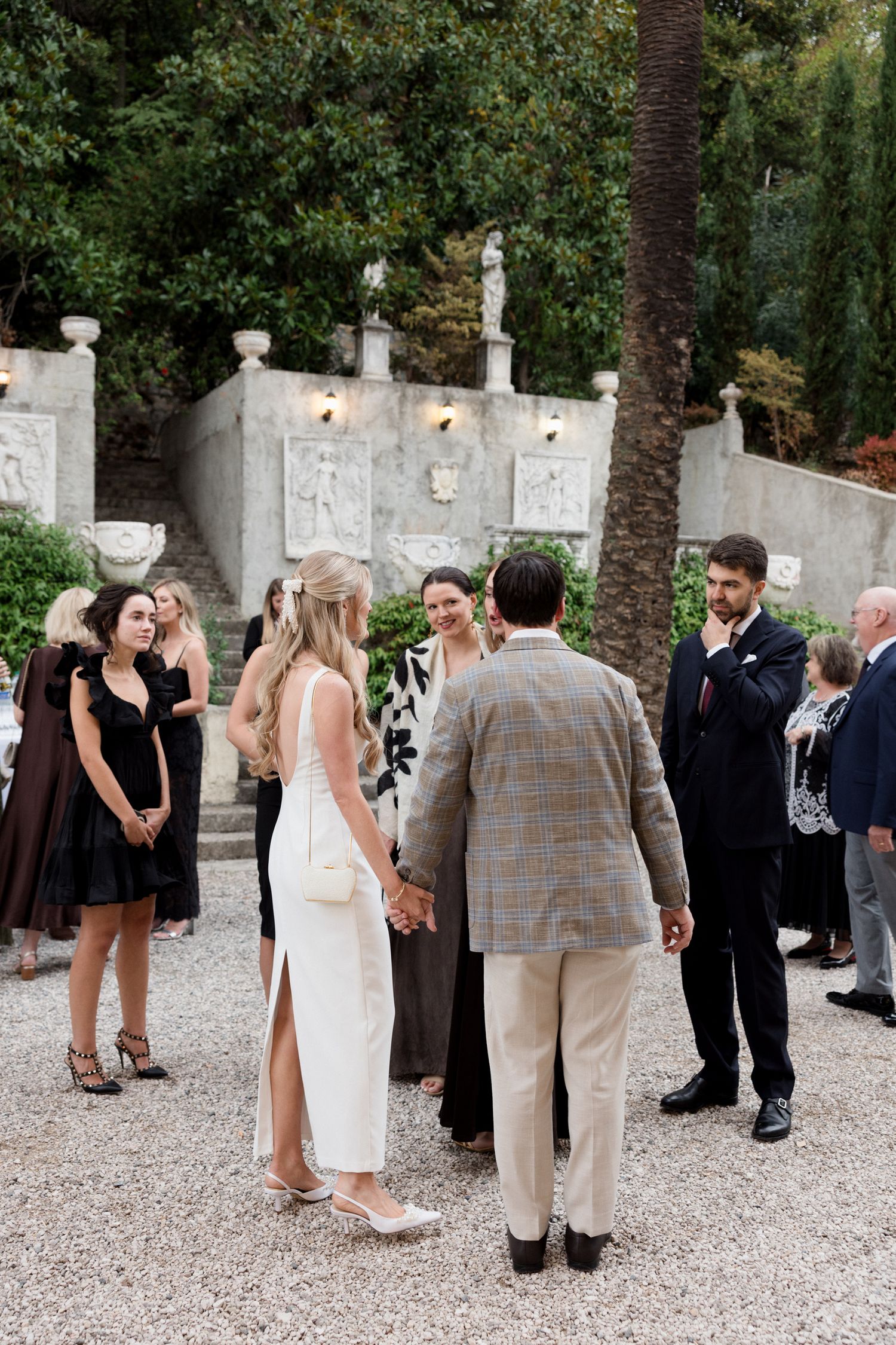 Wedding guests gather and mingle in an elegant outdoor courtyard with white stone walls and palm trees.