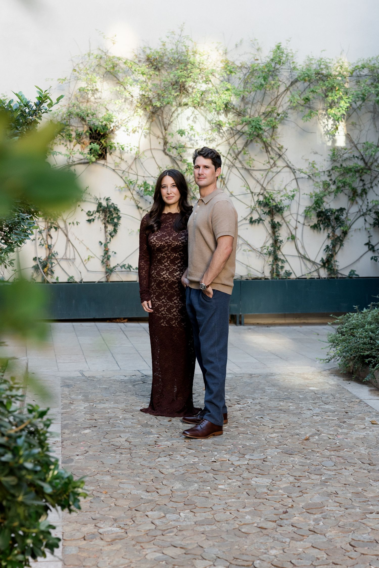 A couple stands together on a stone patio with climbing vines and greenery adorning the white wall behind them. Engagement session Paris