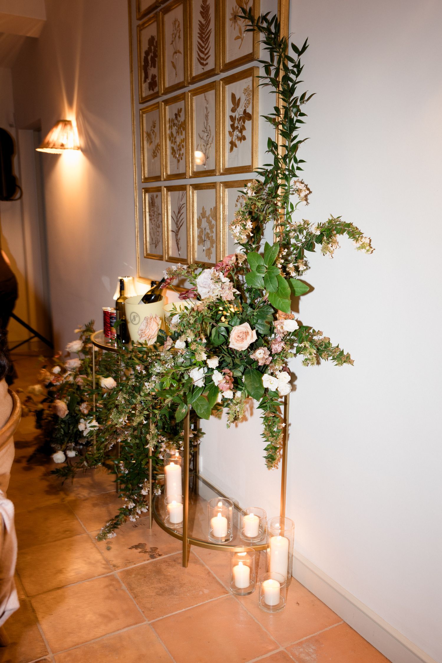 Elegant floral arrangement with pink roses and greenery alongside glowing candles against a wall with framed botanical prints.