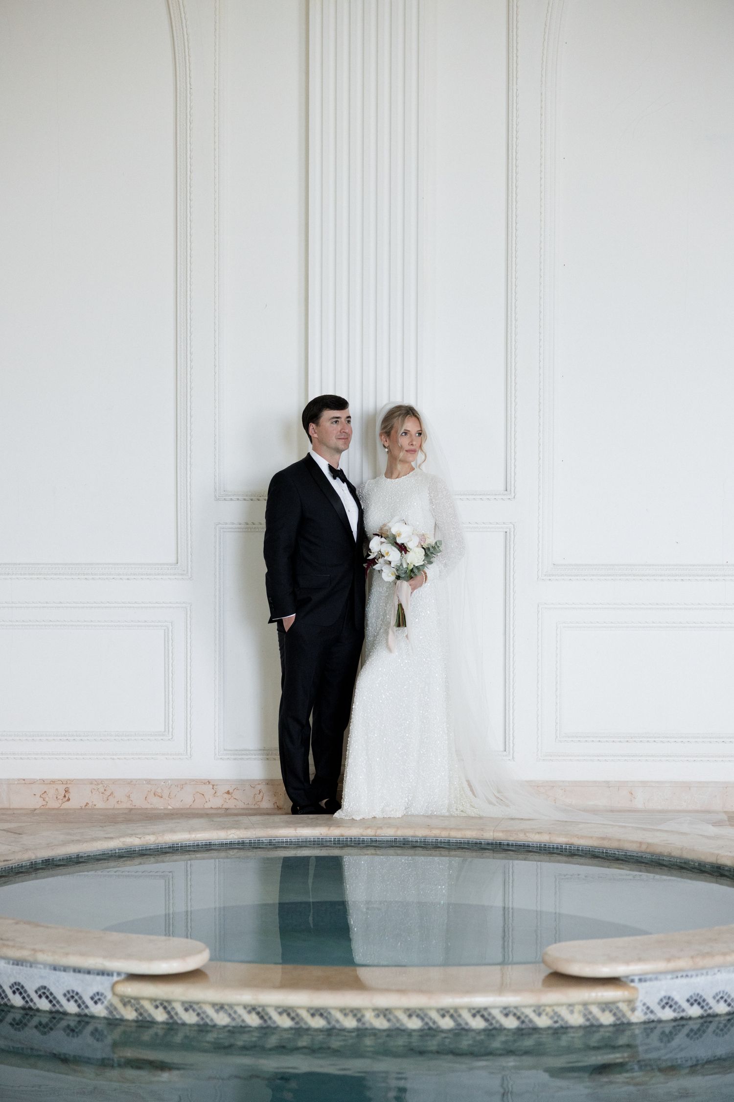 A bride and groom pose against a white wall in an elegant minimalist setting with a pool edge visible.