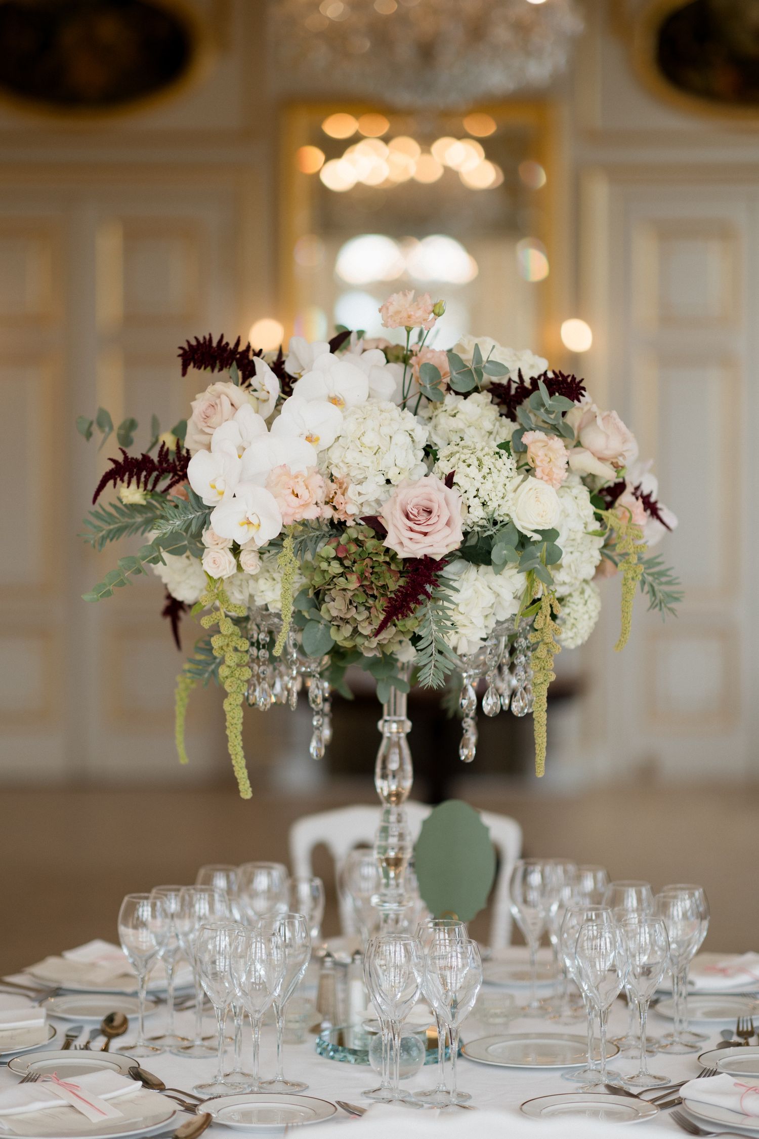 Elegant wedding centerpiece with white and blush roses, orchids and burgundy accents on crystal stand in luxury ballroom setting.