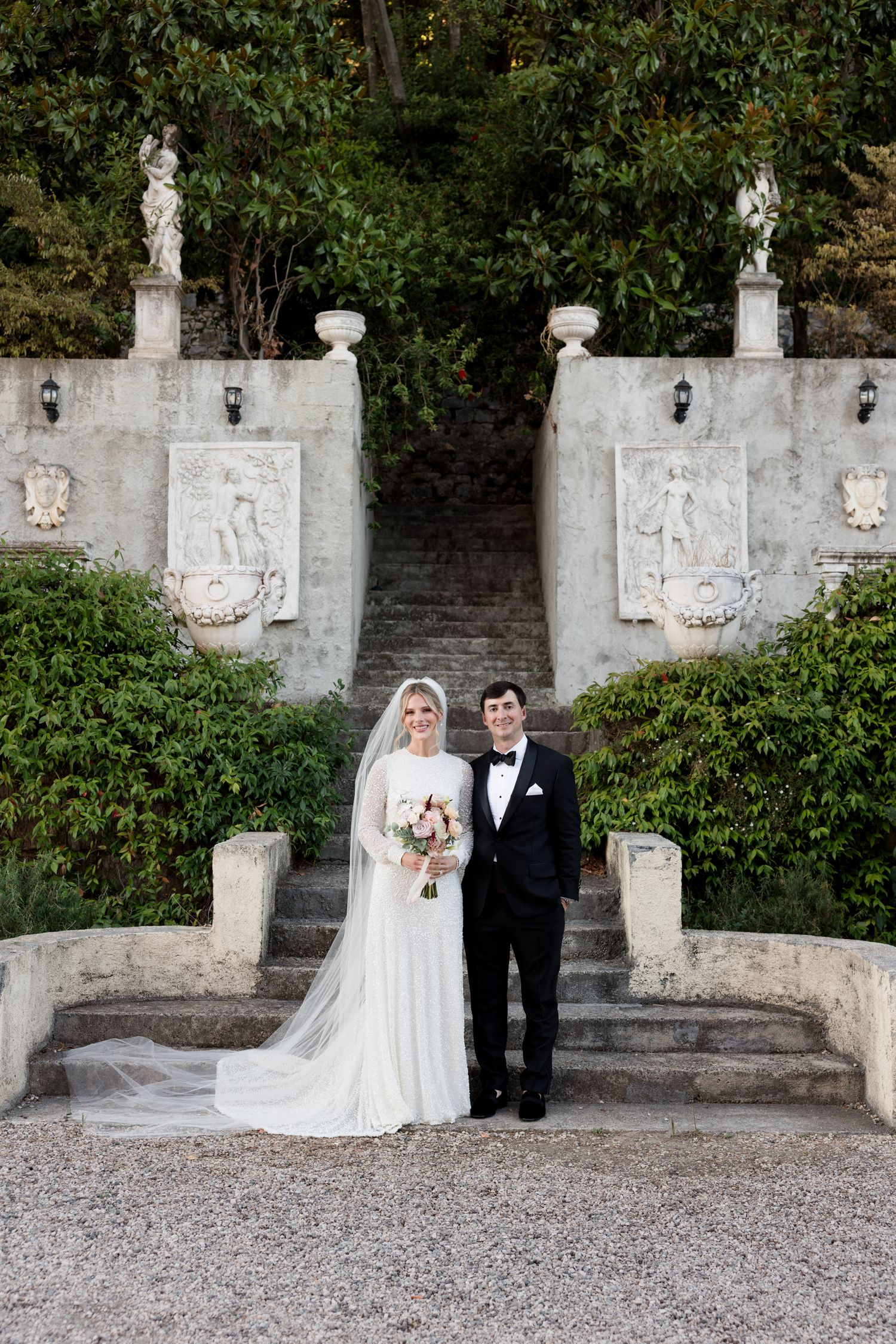 Wedding couple poses in front of decorative white stone wall with carved reliefs and garden steps in elegant outdoor setting.