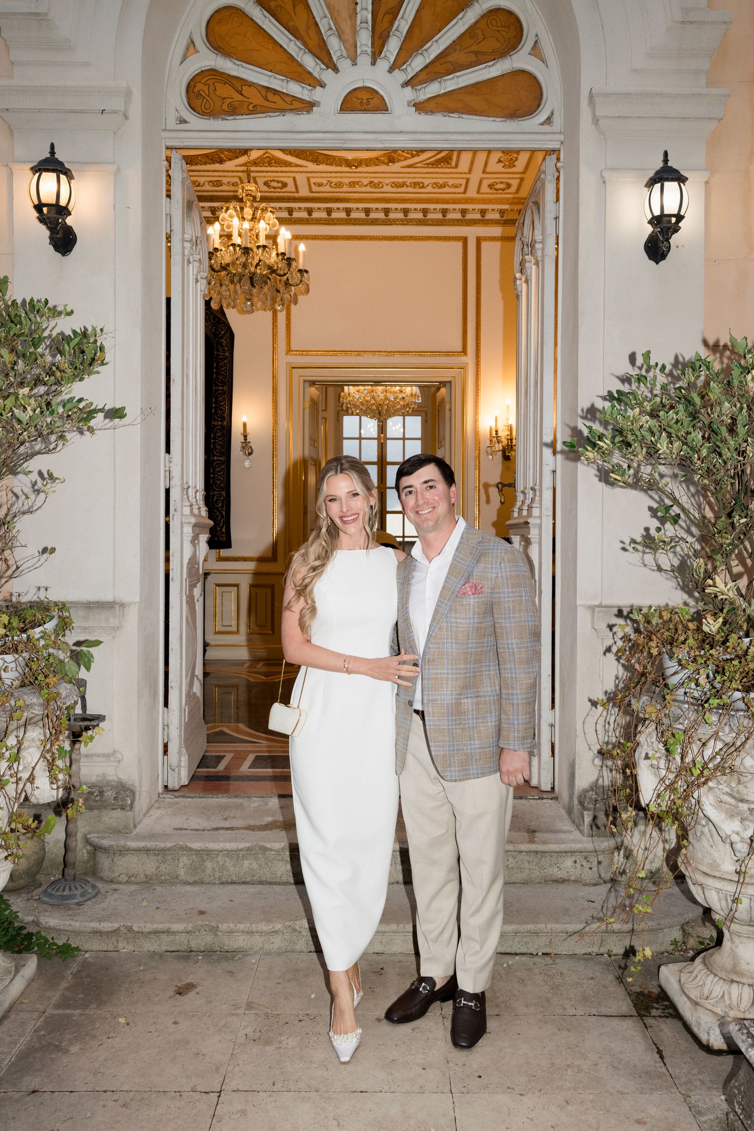 Couple stands together on an elegant porch with white columns, ornate details, and potted olive trees at dusk.