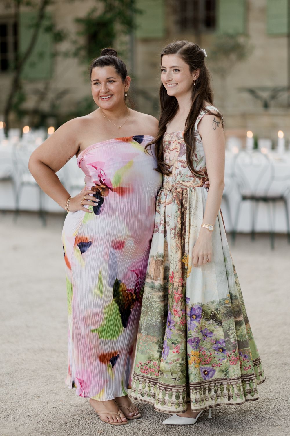 Two people in floral summer dresses stand together on a gravel path at an outdoor event, showcasing colorful patterns.