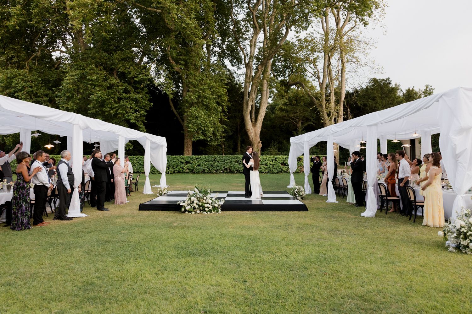 Row of white wedding tents set up on a lush green lawn with trees creating a natural outdoor ceremony space.