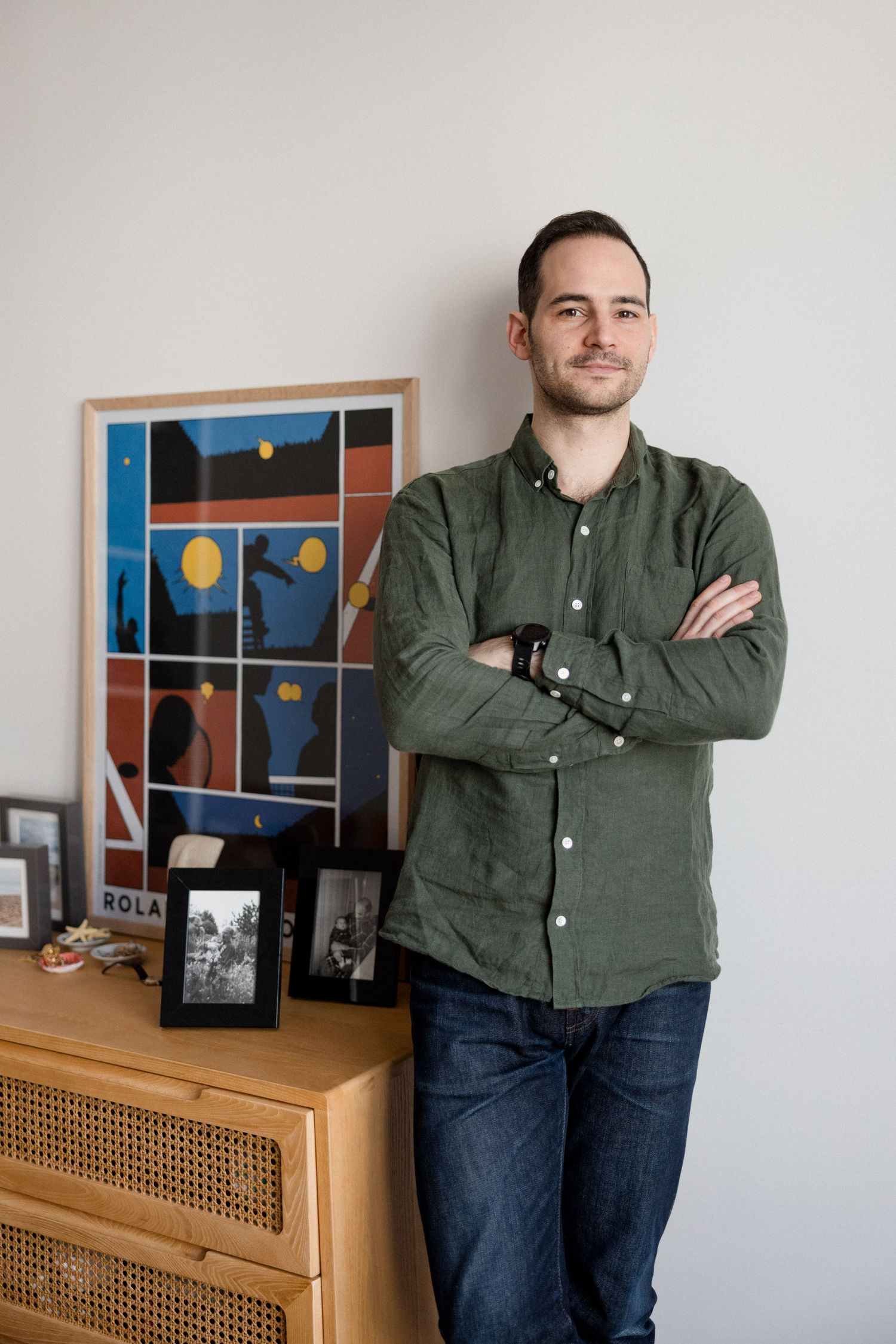 Man in green linen shirt stands arms crossed beside a dresser with colorful framed art and photos.