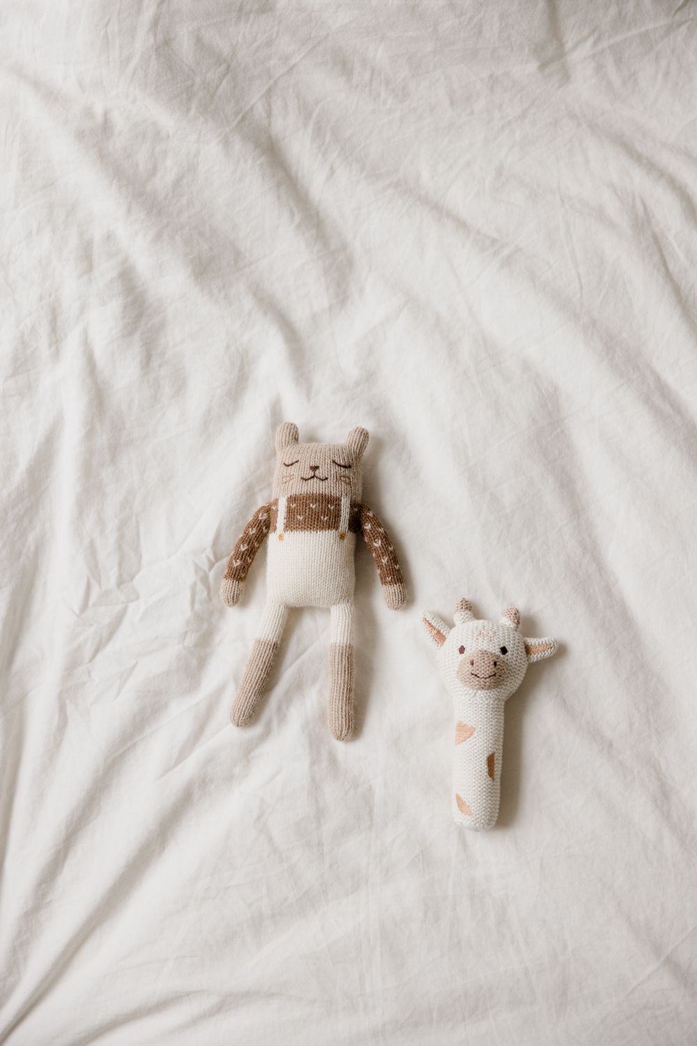 Two soft plush toys, a beige bunny wearing overalls and a small spotted giraffe rattle, placed on white bedding.