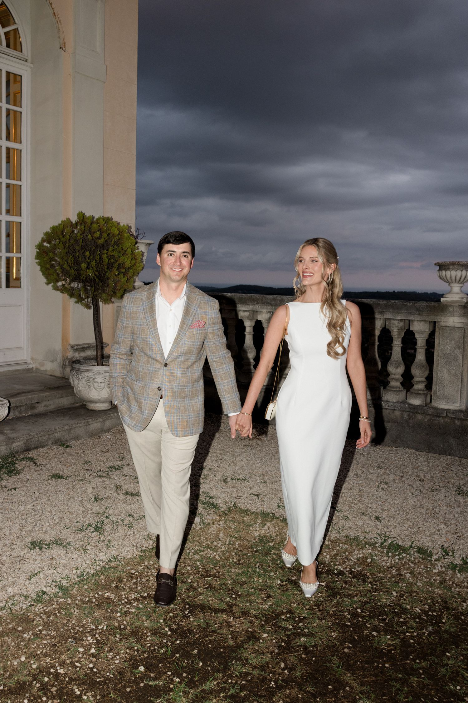 A couple in wedding attire walks together through a dramatic outdoor setting with stormy clouds overhead. destination wedding france