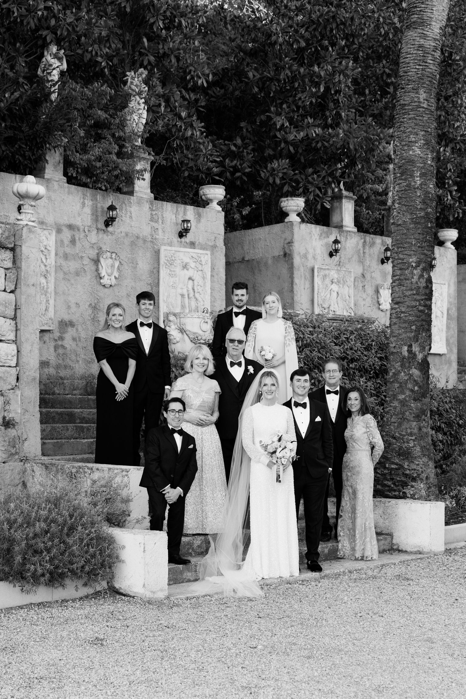 A black and white wedding party poses on elegant stone steps with decorative pillars and landscaping in the background.