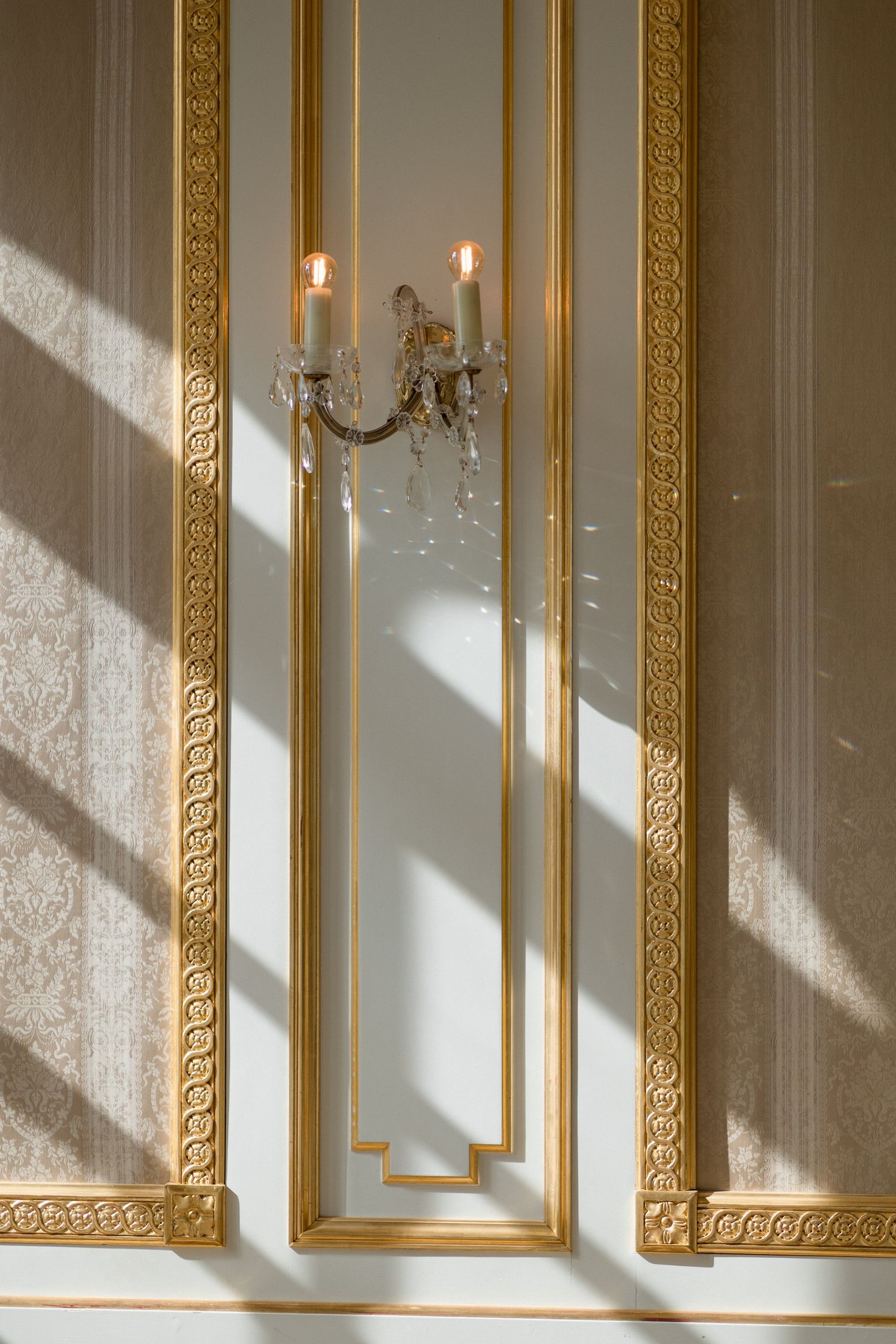 Elegant white wall with ornate gold-framed mirrors and crystal candle sconces casting dramatic diagonal shadows.