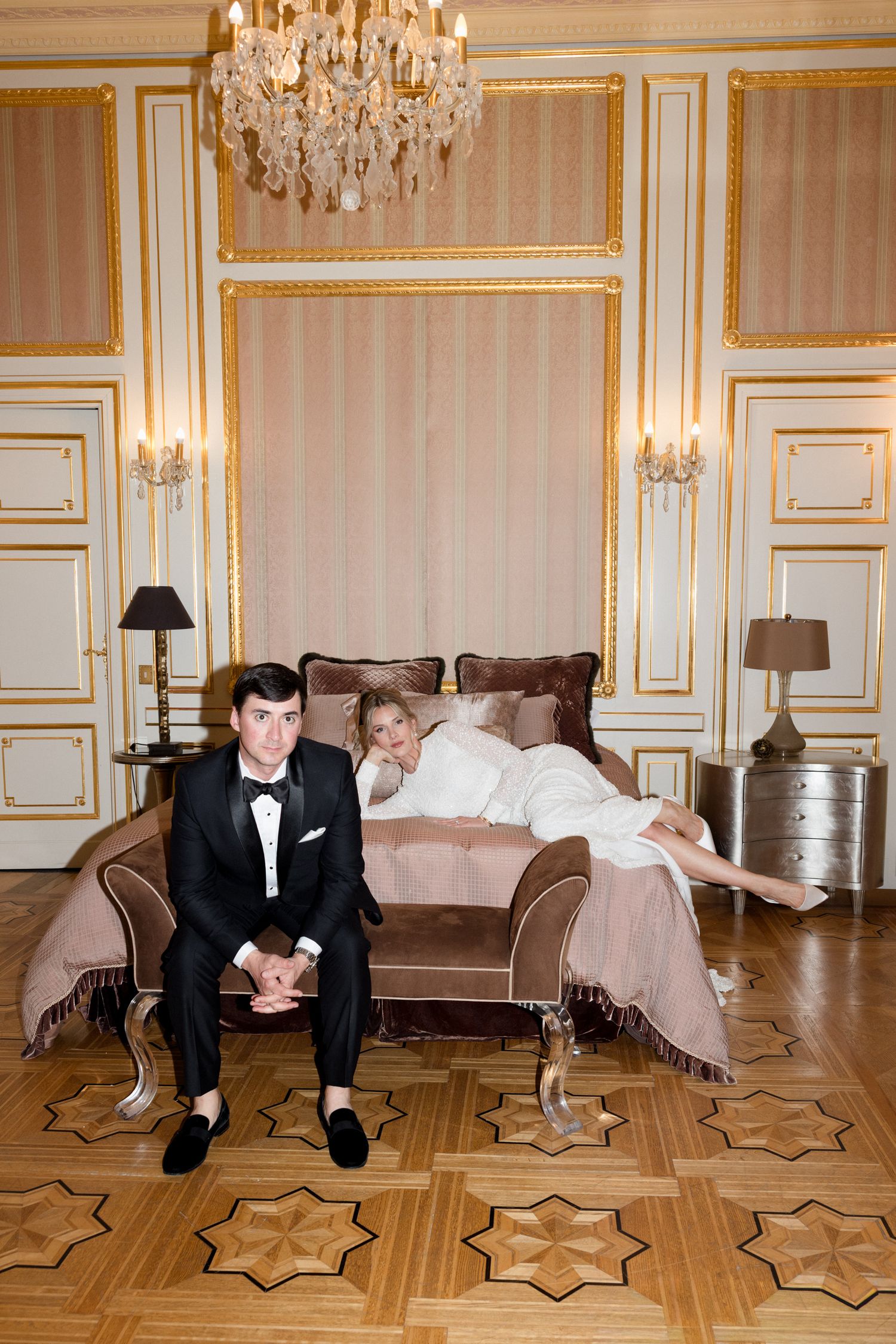 Wedding couple poses playfully on ornate parquet floor beneath crystal chandelier in luxury room.