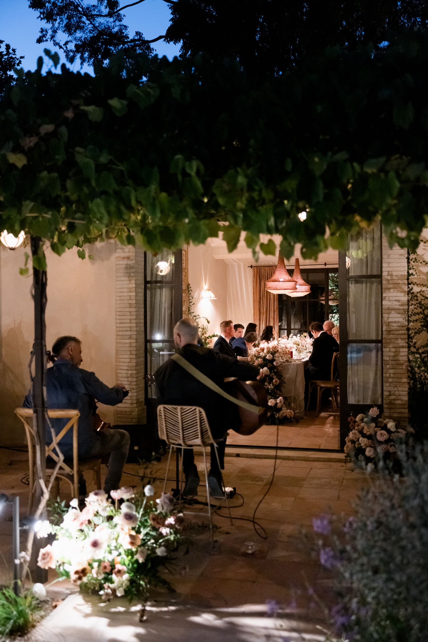 People gather on a dimly lit outdoor patio with string lights and flowers creating an intimate evening ambiance.