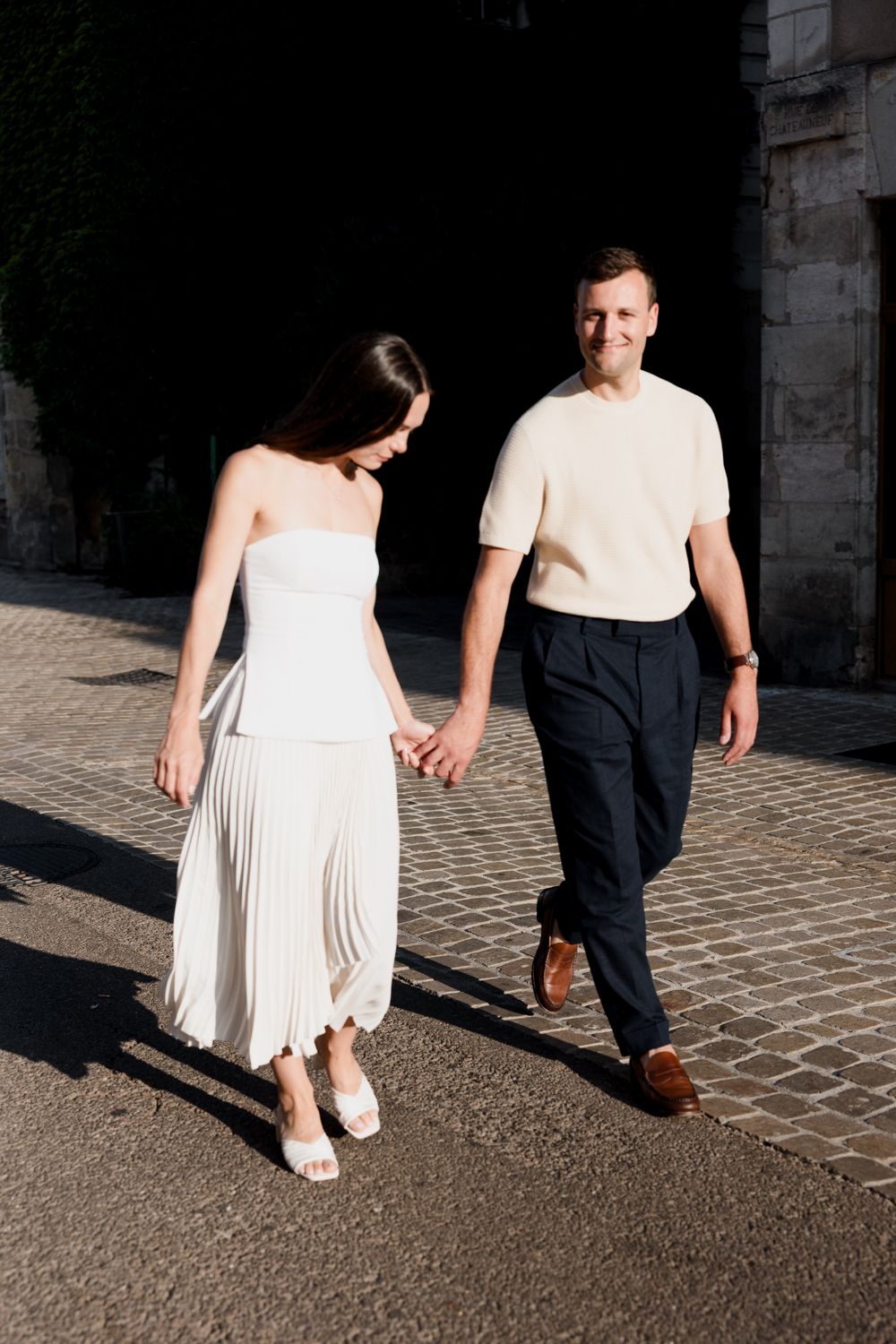 Cropped view of couple walking hand in hand, bride in white dress and groom in dark suit casting shadows on cobblestone path.