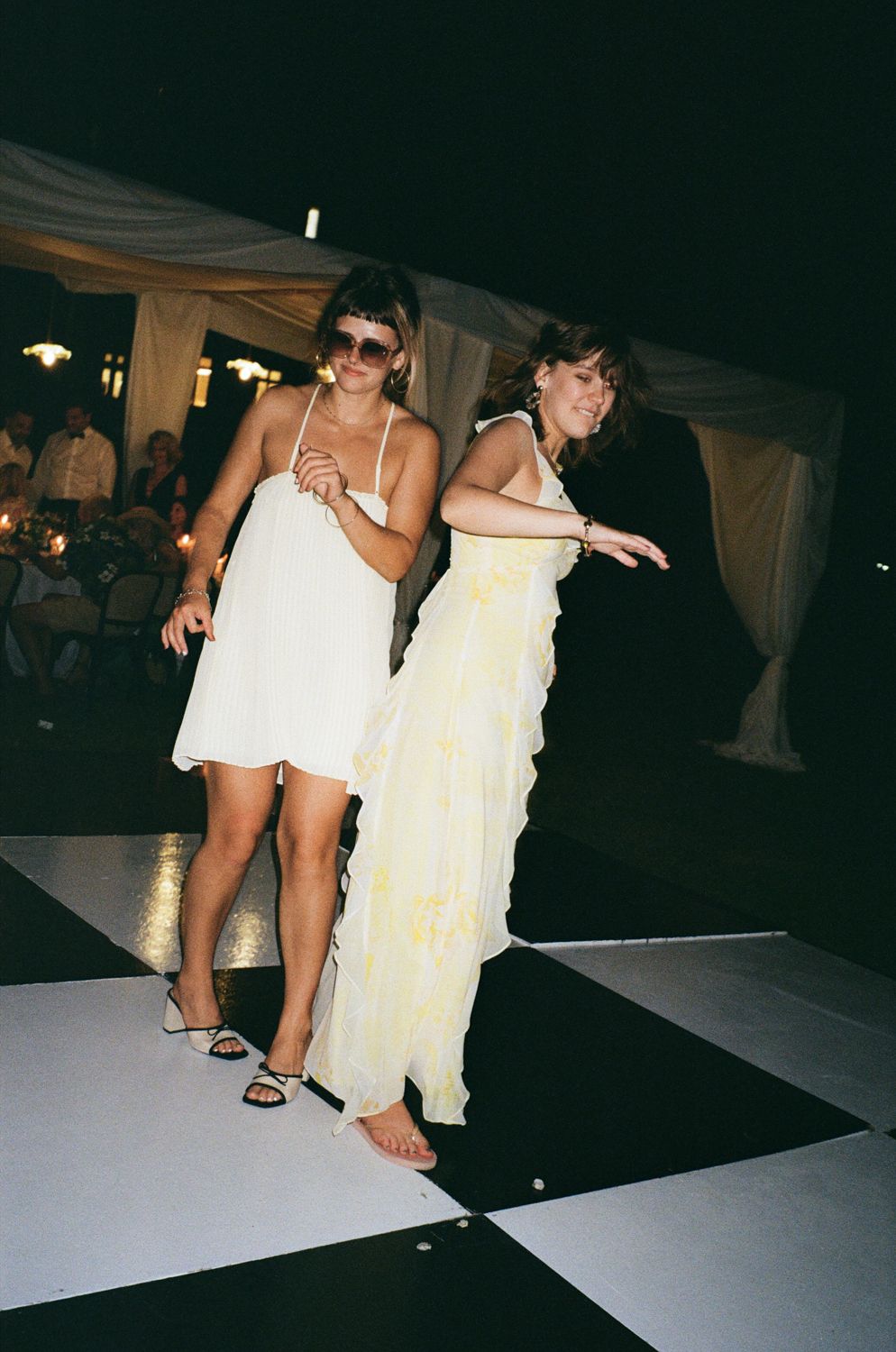 Two people wearing white dresses dance on a black surface at a nighttime event. Wedding on film