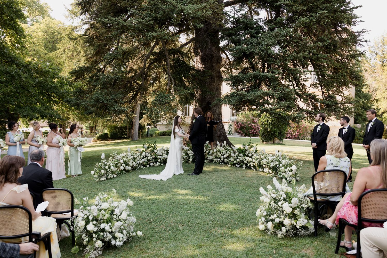 Outdoor wedding ceremony setup under tall trees with white floral arrangements along aisle and white chairs for guests.