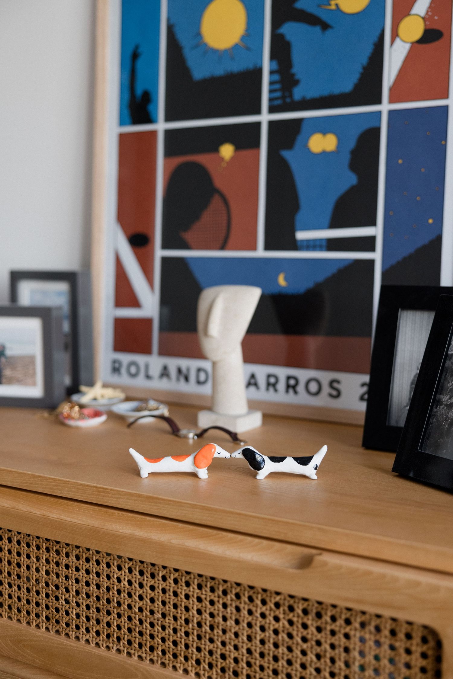 Two ceramic dachshund figurines on a wooden cabinet beside a Roland Garros poster and framed photos.
