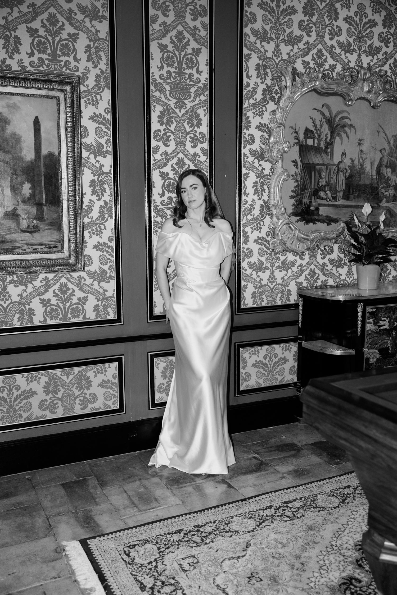 A woman in an elegant white satin gown poses in an ornate room with damask wallpaper and framed artwork. Paris wedding photographer