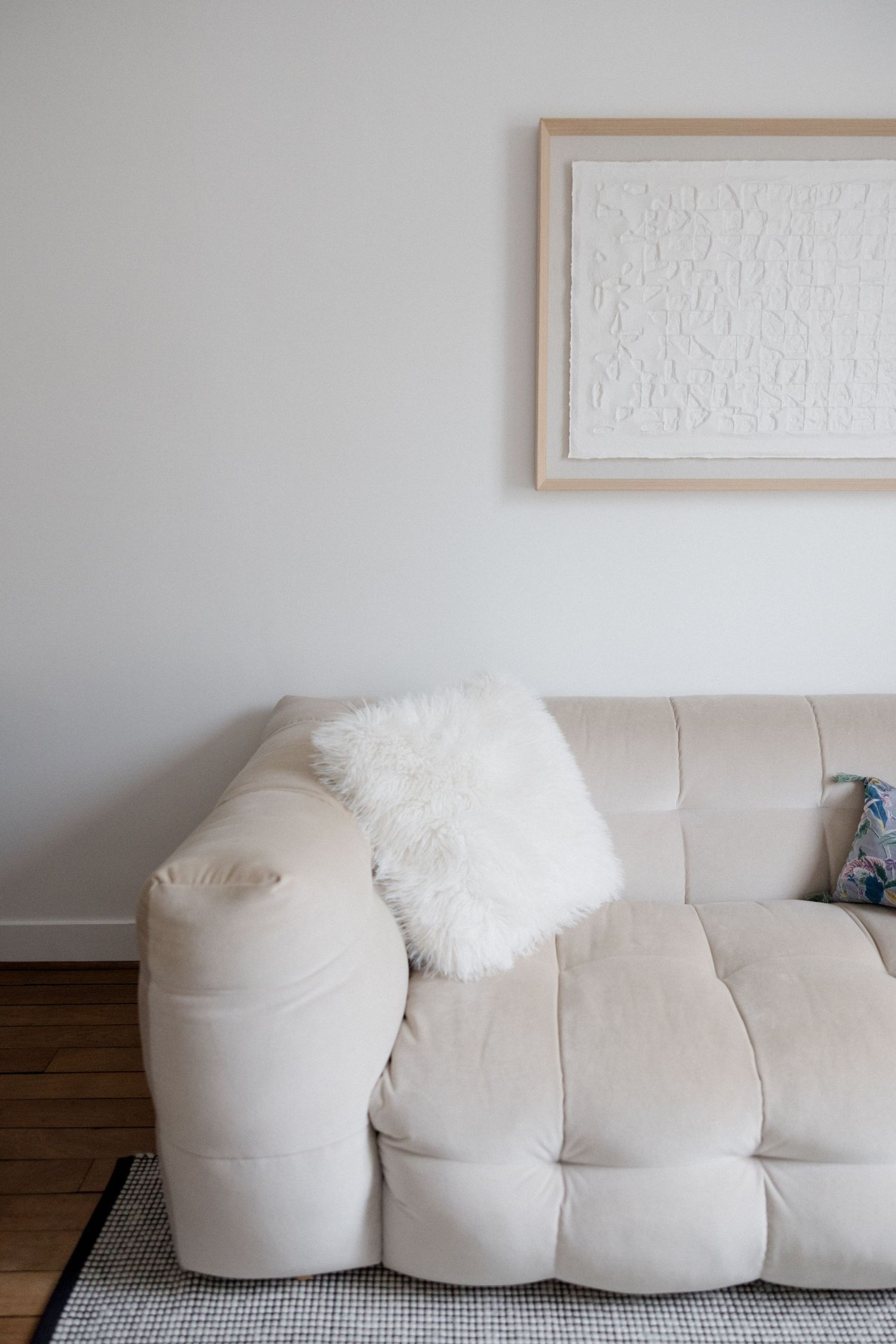 Minimalist living room with a cream tufted sofa, fluffy white pillow, and abstract white artwork in a natural wood frame.