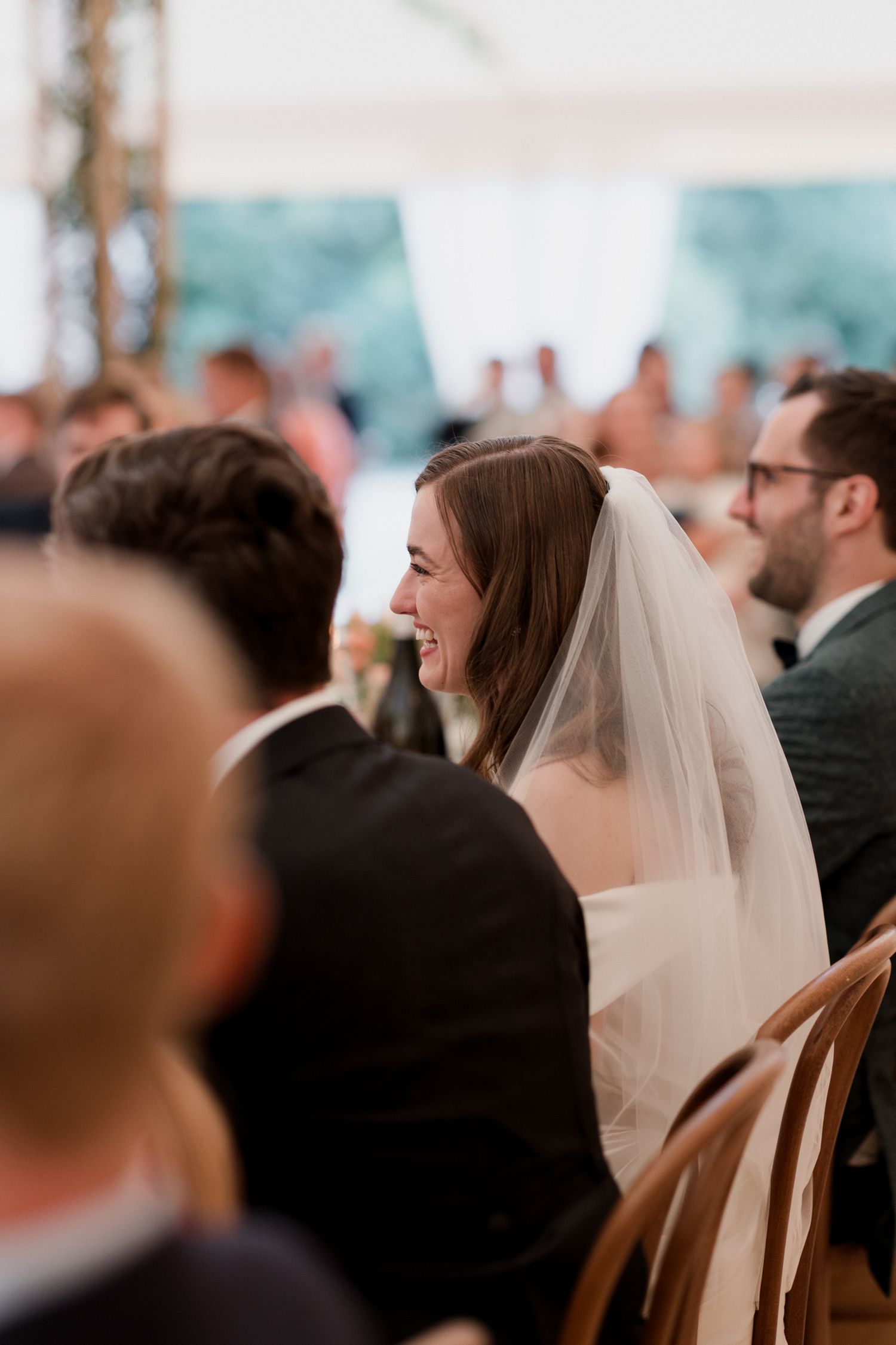 Wedding ceremony guests seated under white tent with natural lighting filtering through.