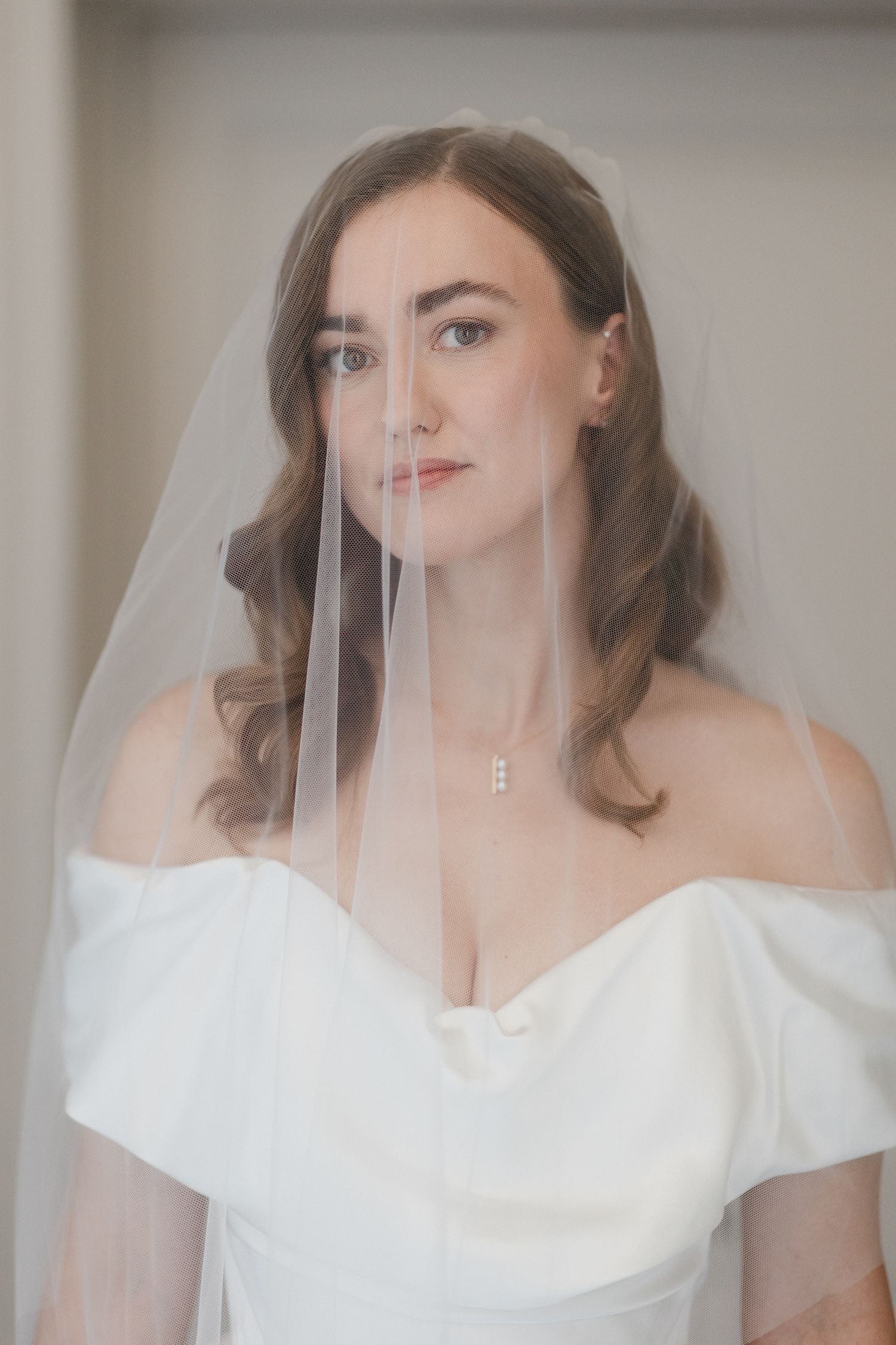 A bride in a classic off-shoulder white wedding dress poses behind a delicate veil with soft natural makeup and wavy hair.