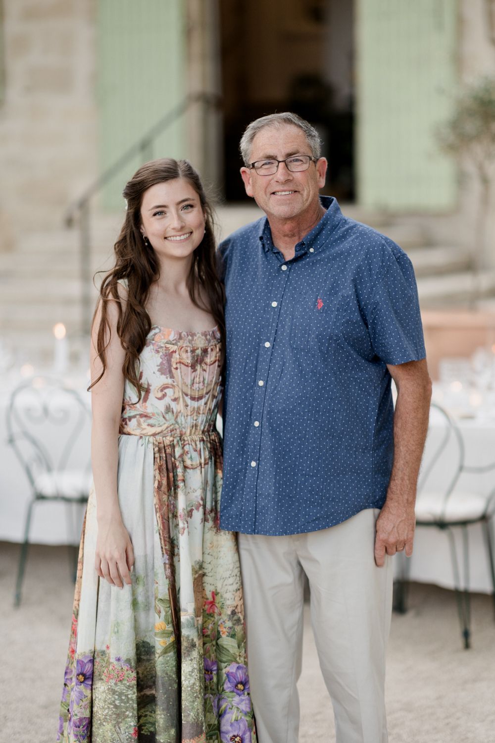 A young woman in a floral maxi dress poses outdoors with an older man wearing a blue button-down shirt and khakis.