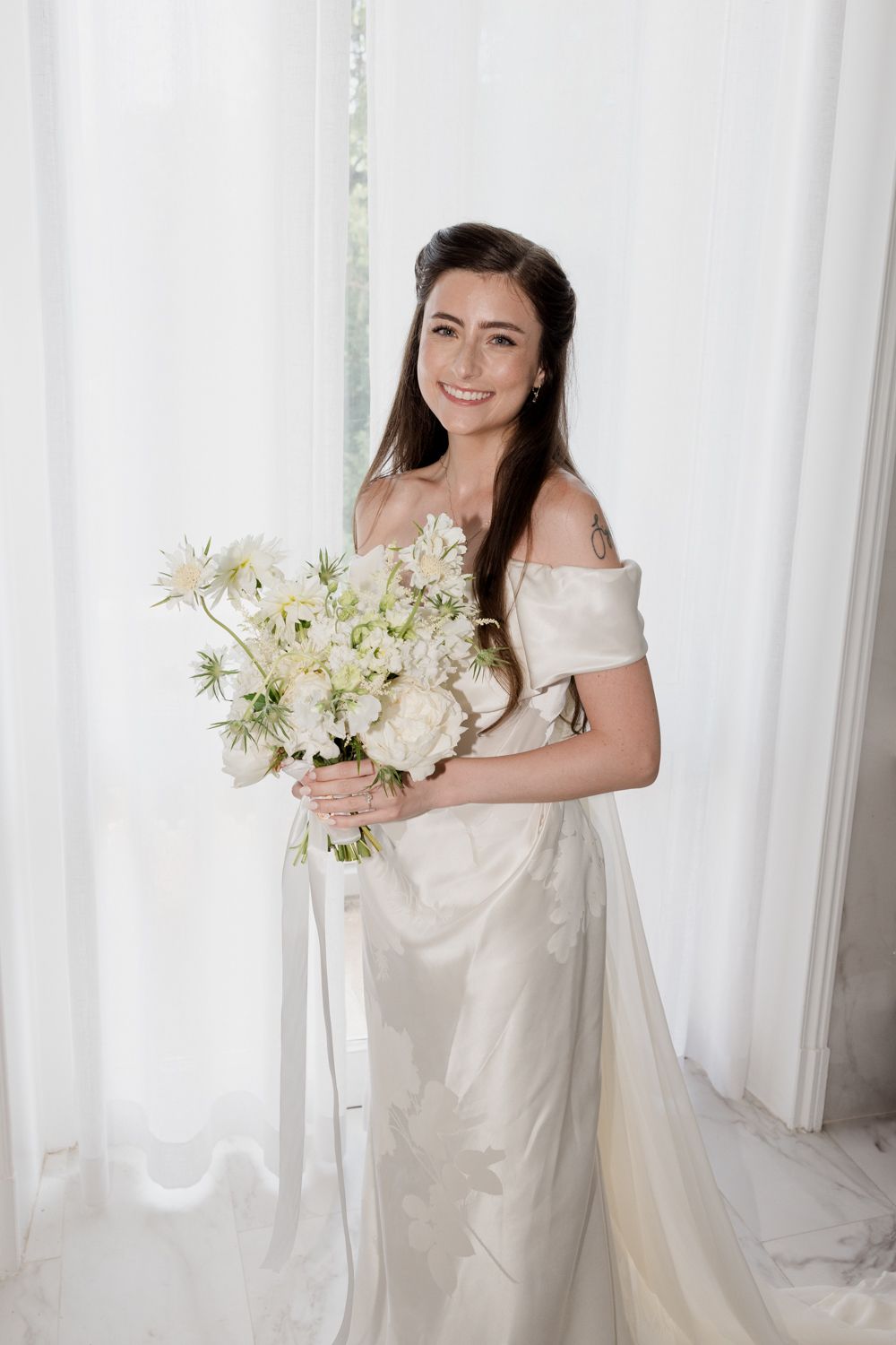 A bridal portrait in white showing elegant poses against sheer curtains while holding a bouquet of white flowers. Photographe mariage Tours