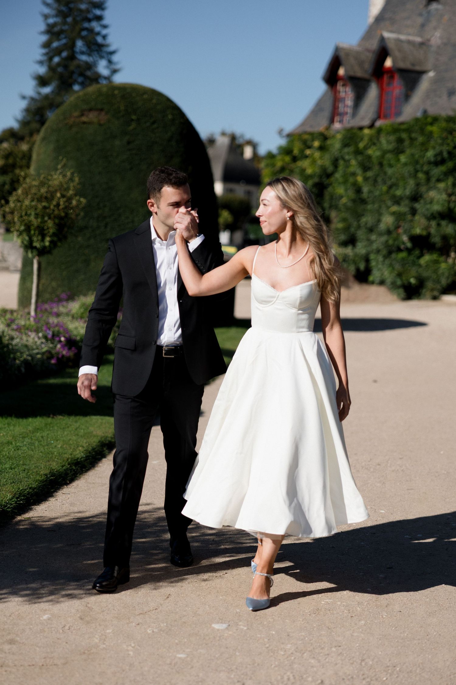 Bride in white tea-length dress and groom in black suit walk together in a romantic French château garden.