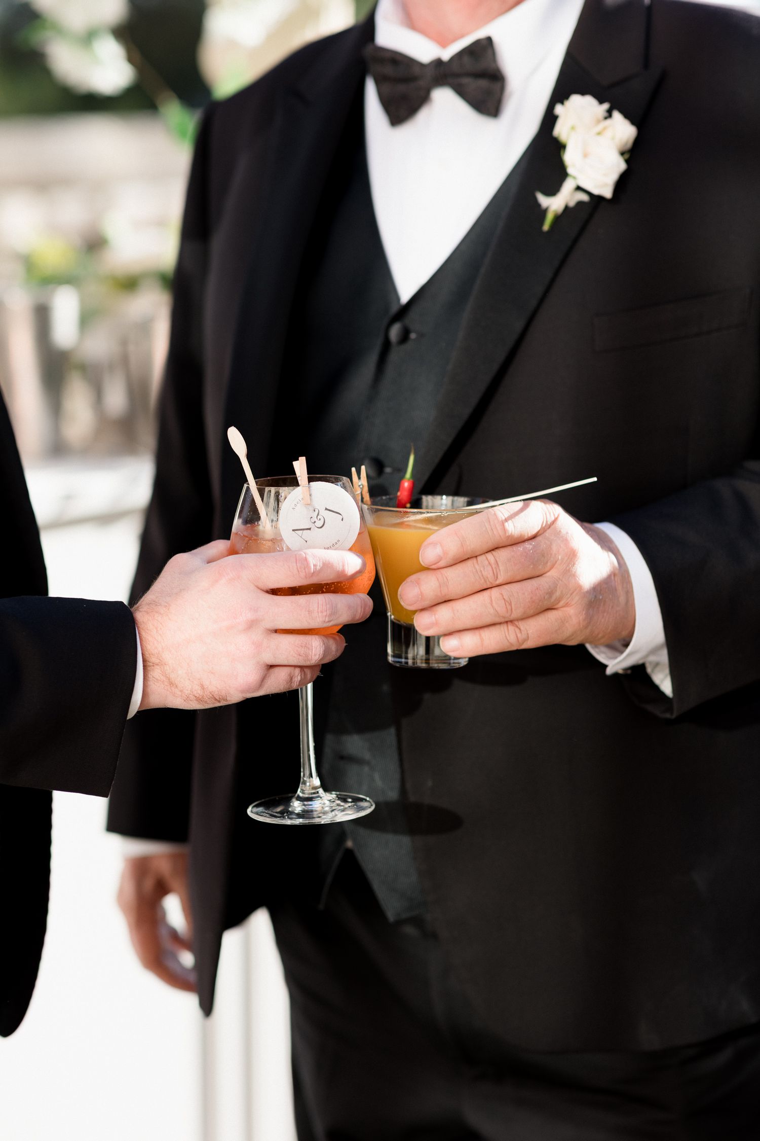 Close-up view of hands holding cocktail glasses during a formal event with black tuxedos and bow ties. Photographe mariage france grasse