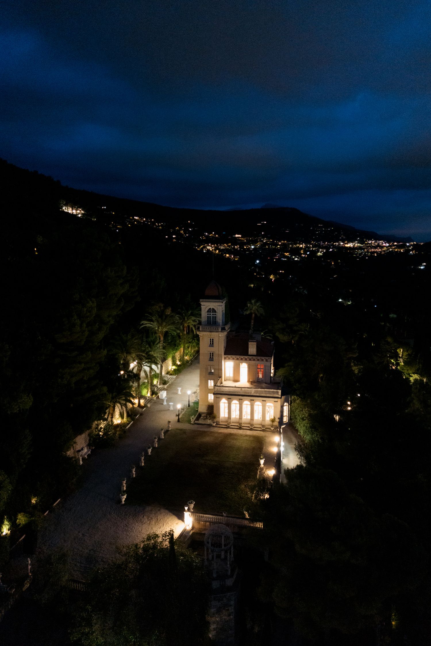 Illuminated church on hillside at night with twinkling city lights in background and dramatic dark blue sky.