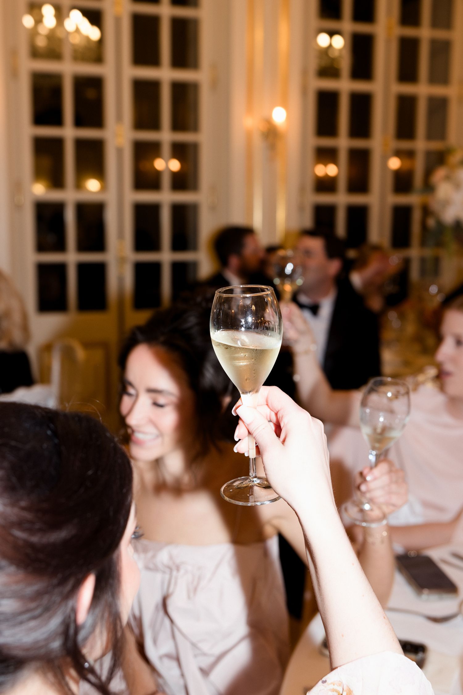 People raise champagne glasses in a toast at an elegant indoor celebration with warm lighting and white decor. Mariage grasse