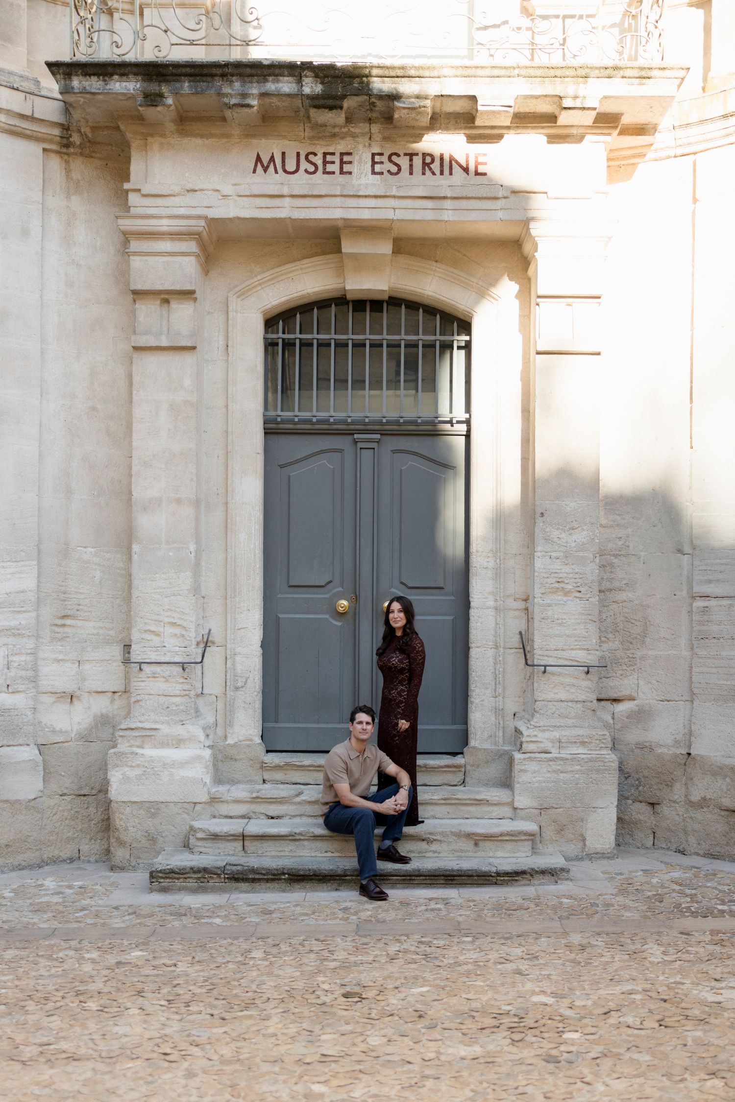 Stone facade of the Musee Estrine with grey doors and visitors sitting on steps in Provence, France. Engagement session France