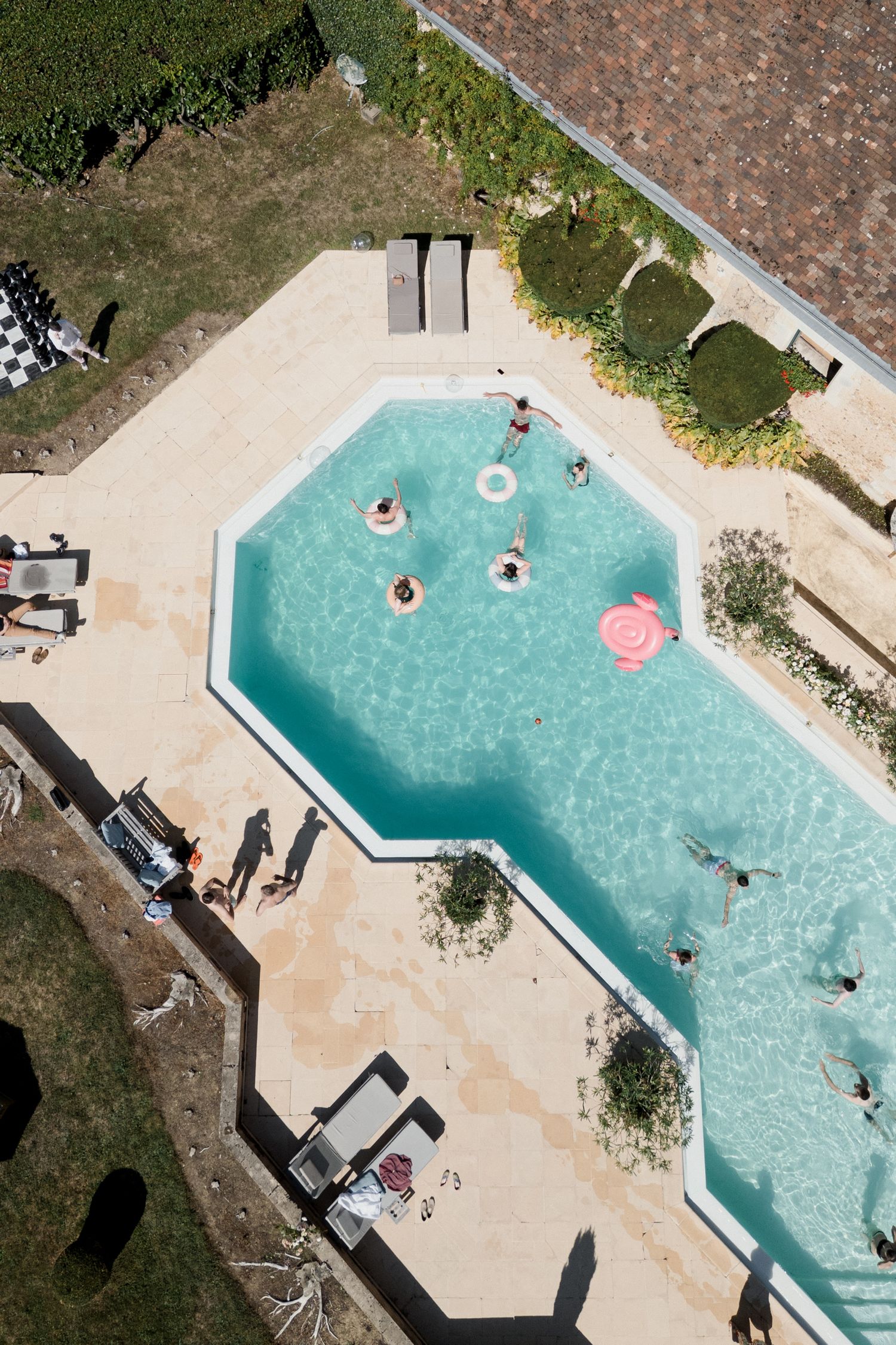 Aerial view of swimmers enjoying a turquoise L-shaped swimming pool surrounded by concrete deck and landscaping. Pool party loire valley