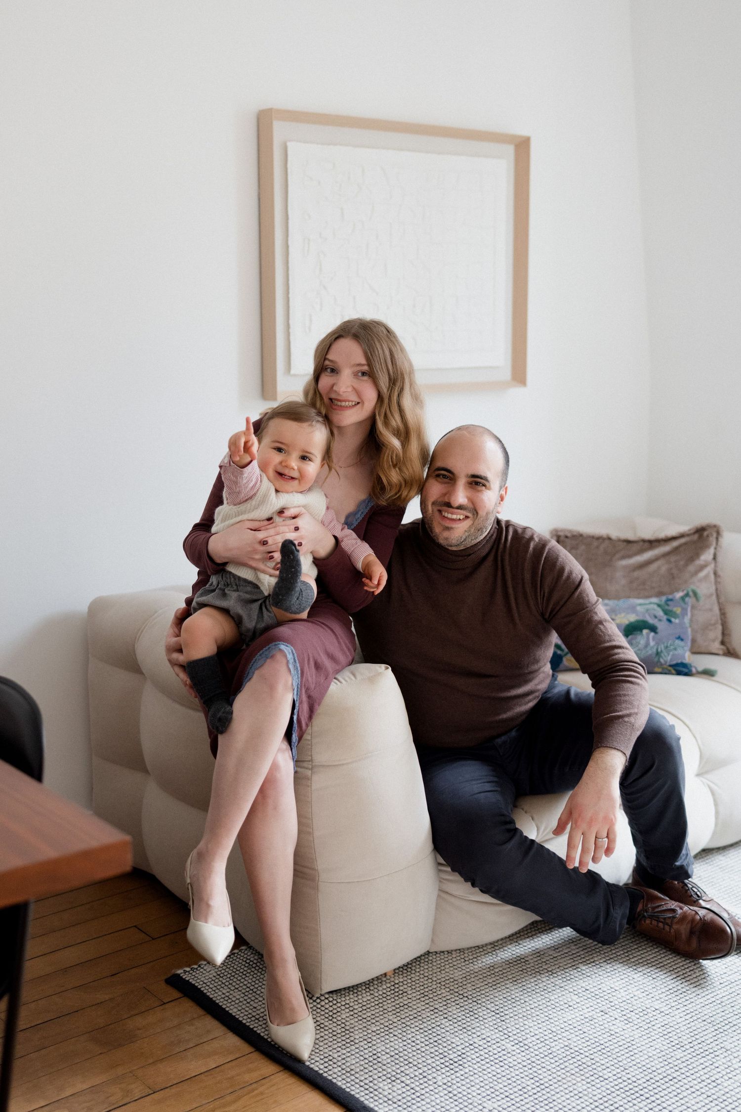 Happy young family with baby sitting on couch in modern living room, smiling at camera.