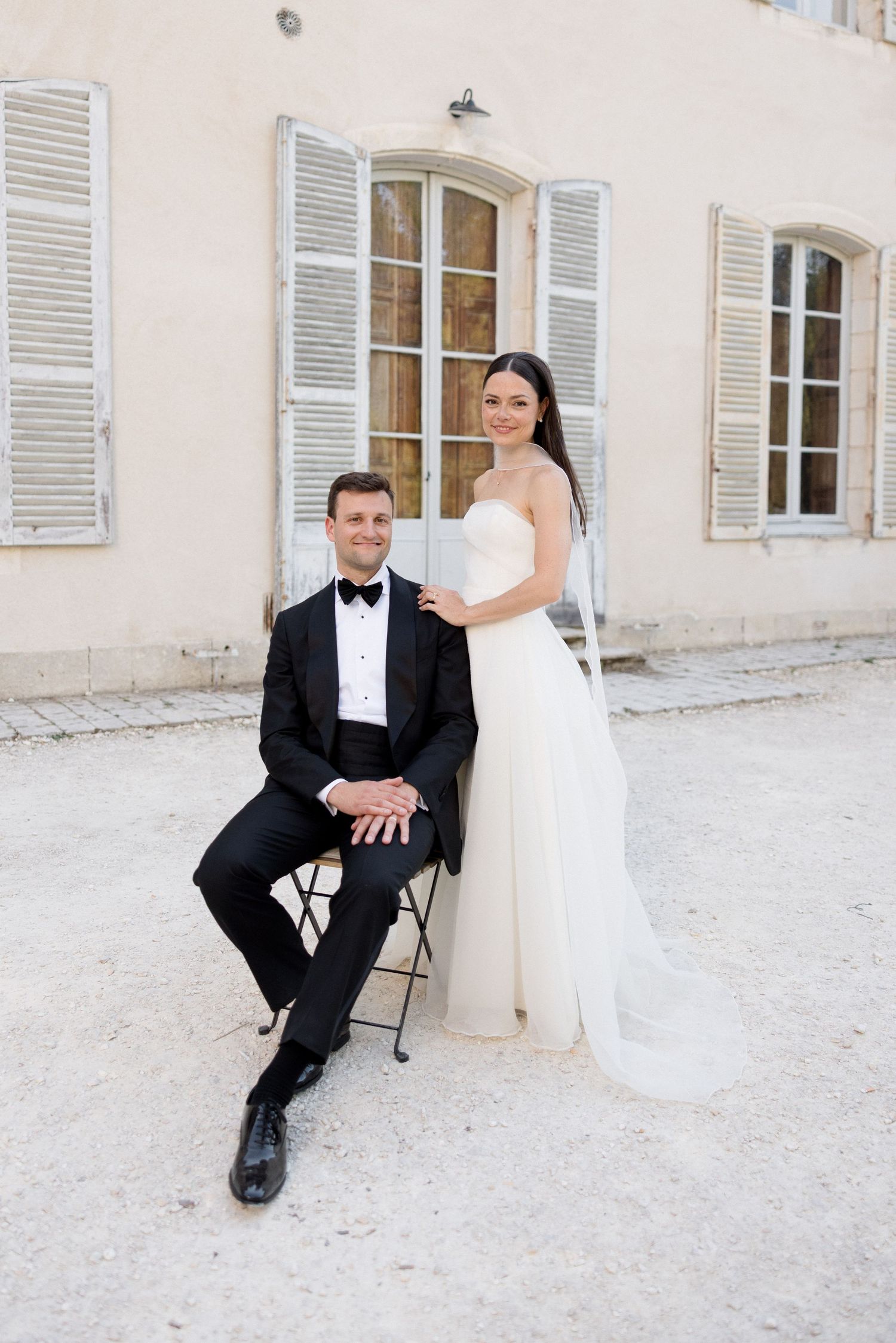 Elegant bride in white gown stands behind groom in black tuxedo seated on chair outside a French chateau.