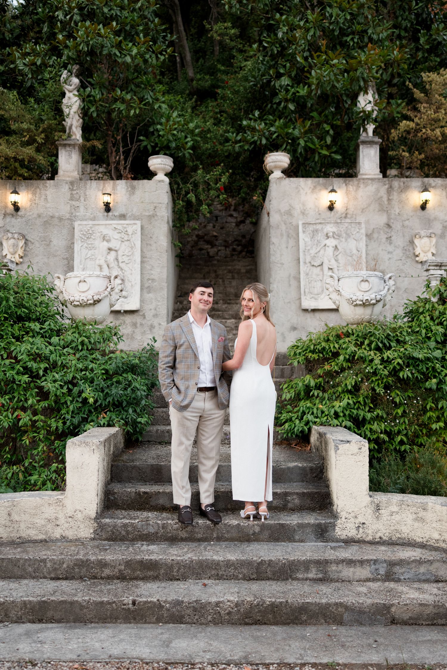 A couple in wedding attire poses against a rustic stone wall with decorative elements in an outdoor garden setting.