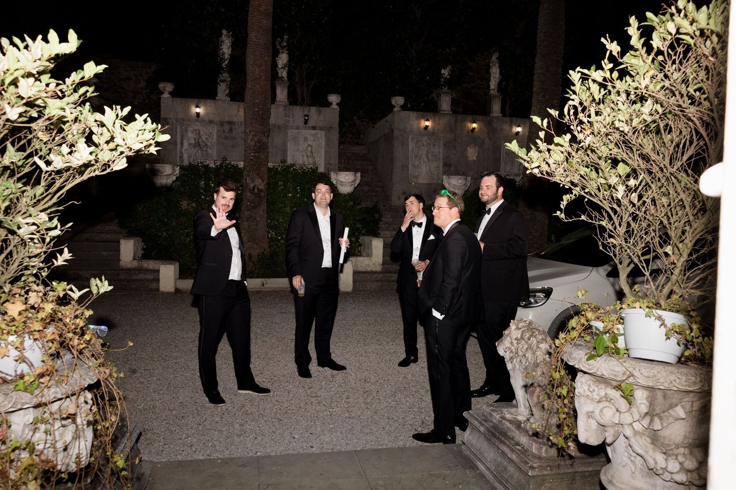 Wedding guests in formal attire gather around elegantly decorated floral arrangements at an evening reception.