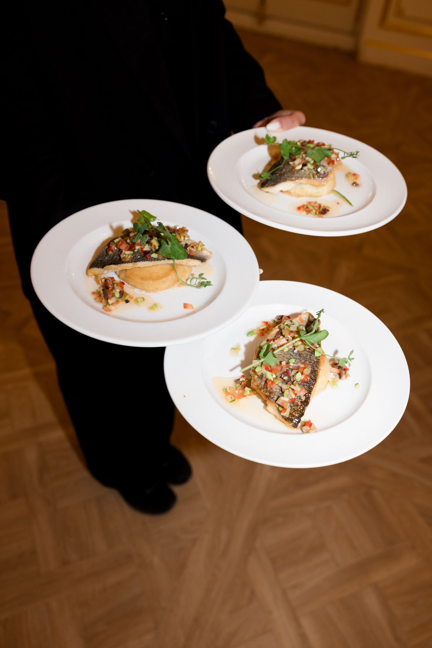 Three white plates with elegantly plated dishes garnished with fresh herbs sit on a dark wooden surface.