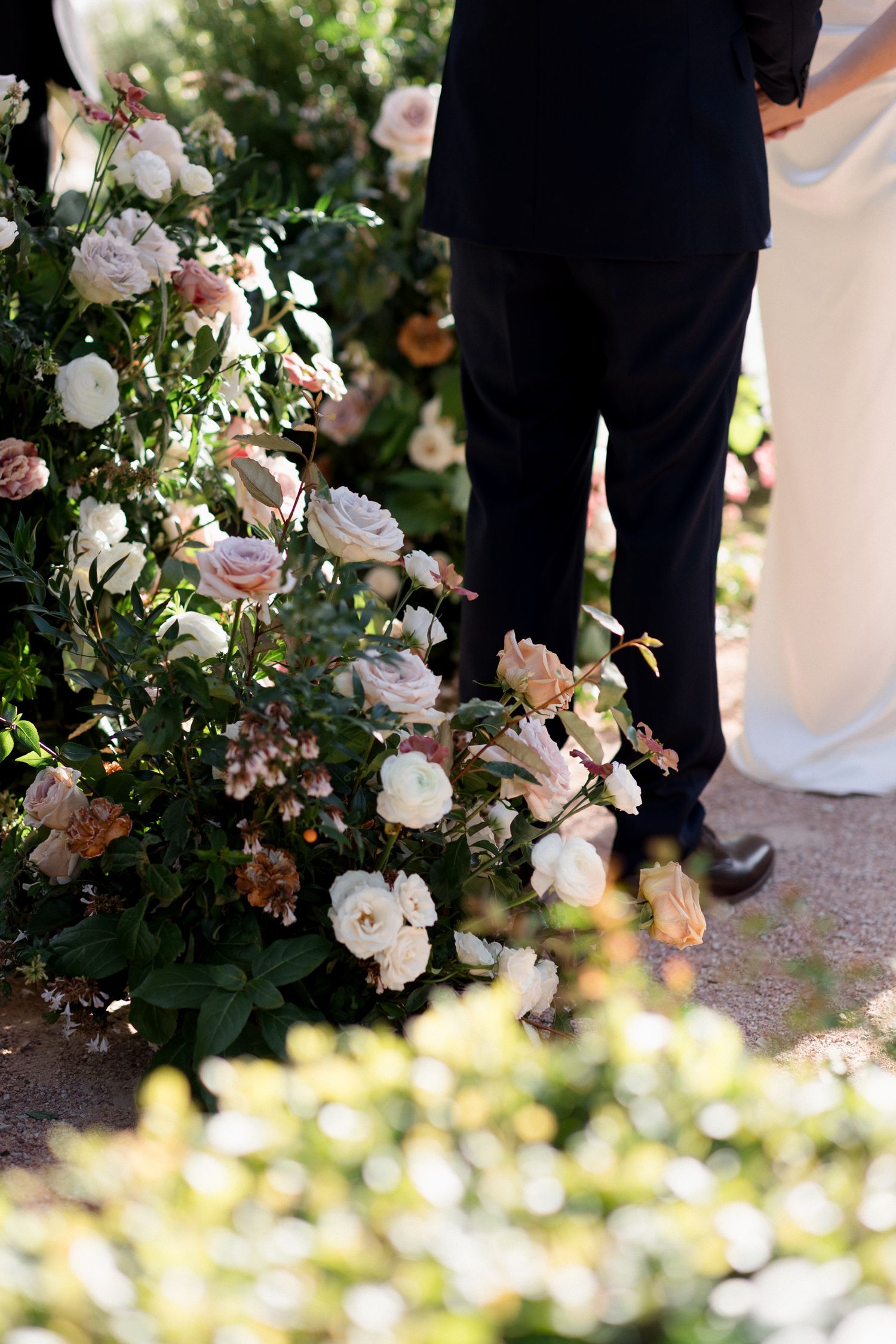 Close-up view of wedding ceremony details showing elegant floral arrangements and formal attire in soft natural lighting.