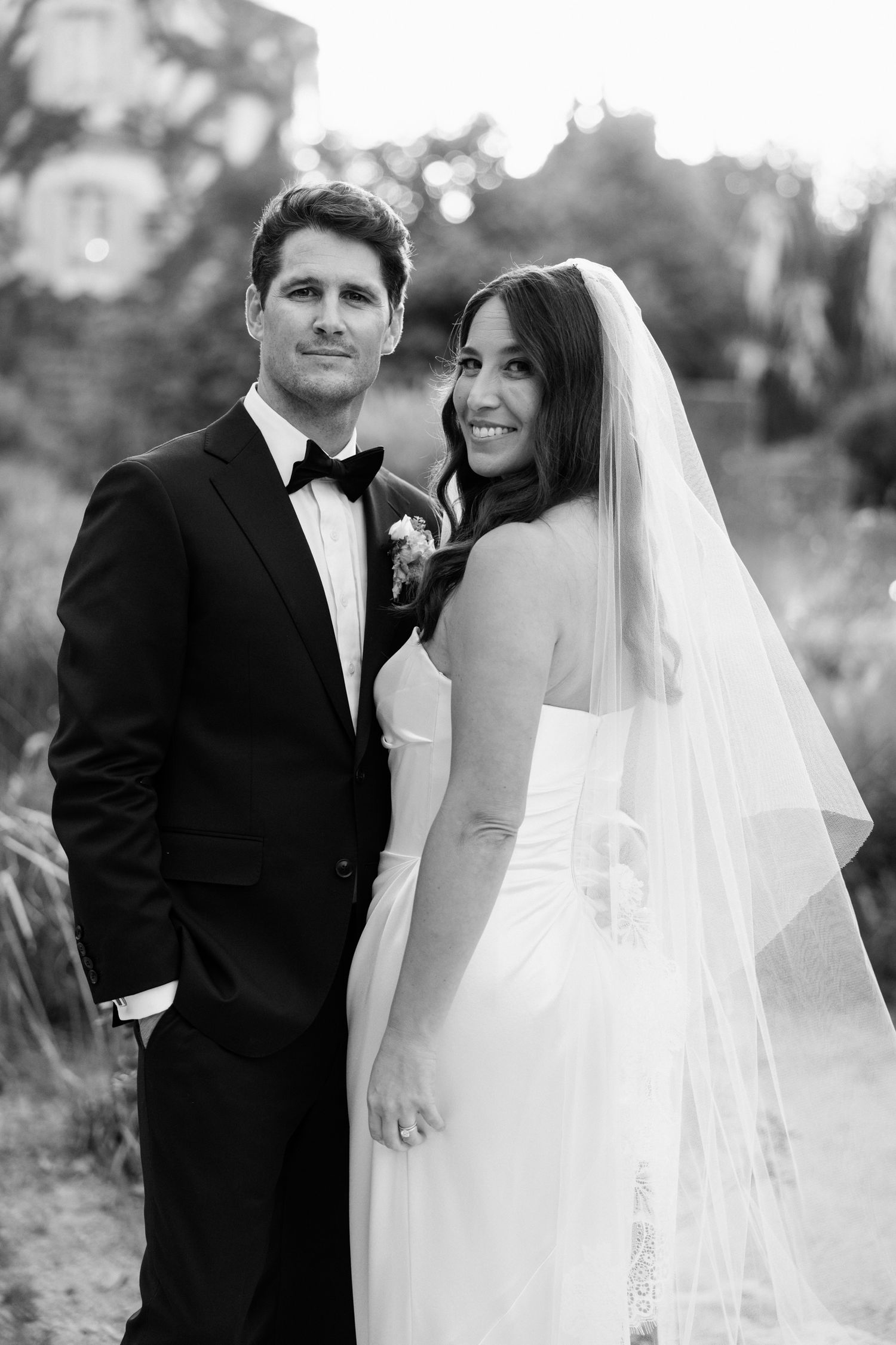 A black and white wedding portrait of a couple outdoors, with the bride in a white gown and veil and groom in a tuxedo.