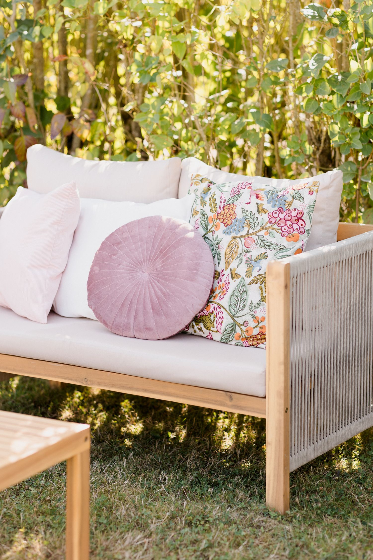 Elegant outdoor wooden daybed with pink and floral throw pillows arranged in a garden setting with natural foliage backdrop. Pool party loir