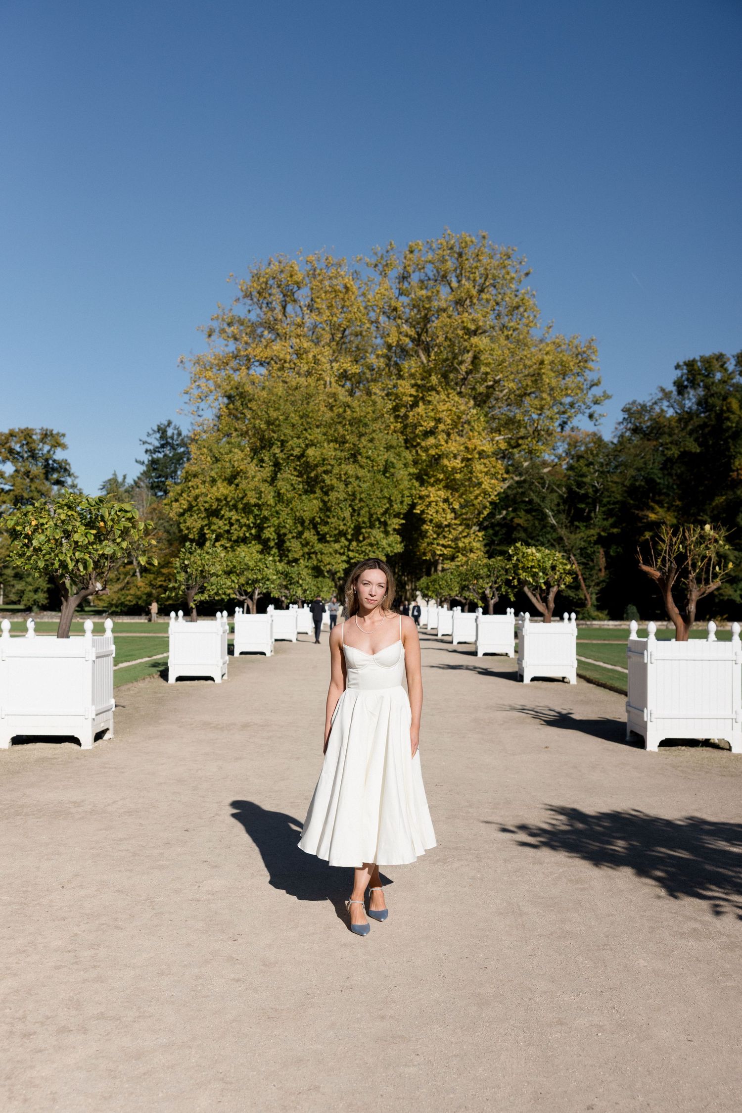 Bride in white midi dress walks along garden path flanked by white planters under clear blue sky.