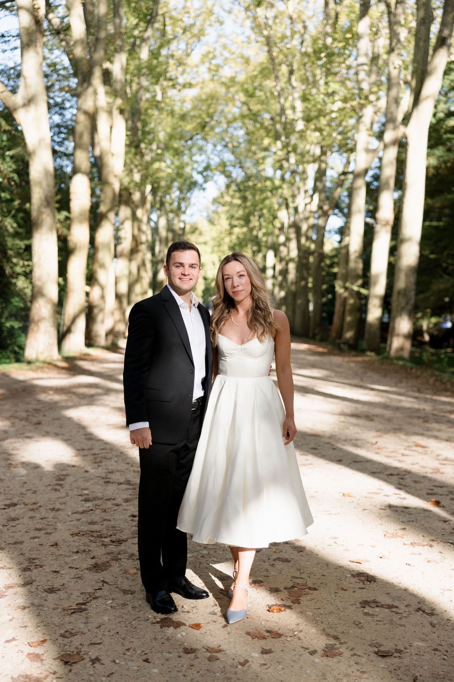 Bride and groom posing on a sunlit tree-lined path, she wearing a white midi dress and he in a black tuxedo.
