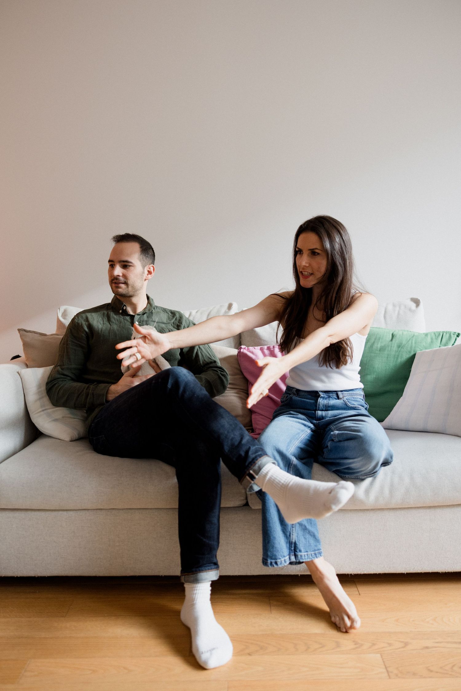 A couple sits on a gray sofa, woman gesturing expressively toward man during a conversation at home.
